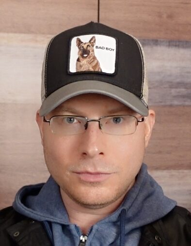 Man wearing glasses, a cap with a dog patch, and a hoodie, looking directly at the camera.