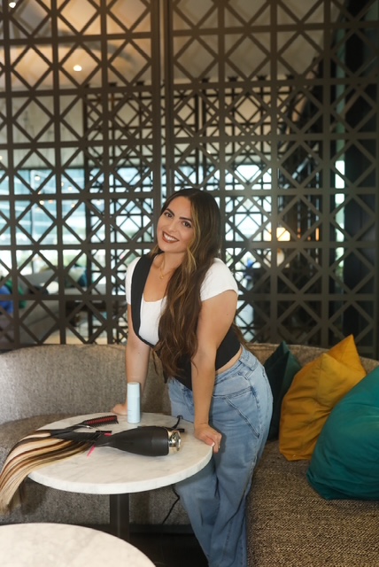 Woman with long hair smiling, standing by a table with a hairbrush and spray bottle, in a cozy seating area.
