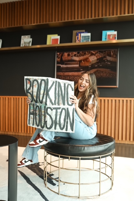Young woman sitting on a round stool holding a sign that says 'Booking Houston' and smiling.