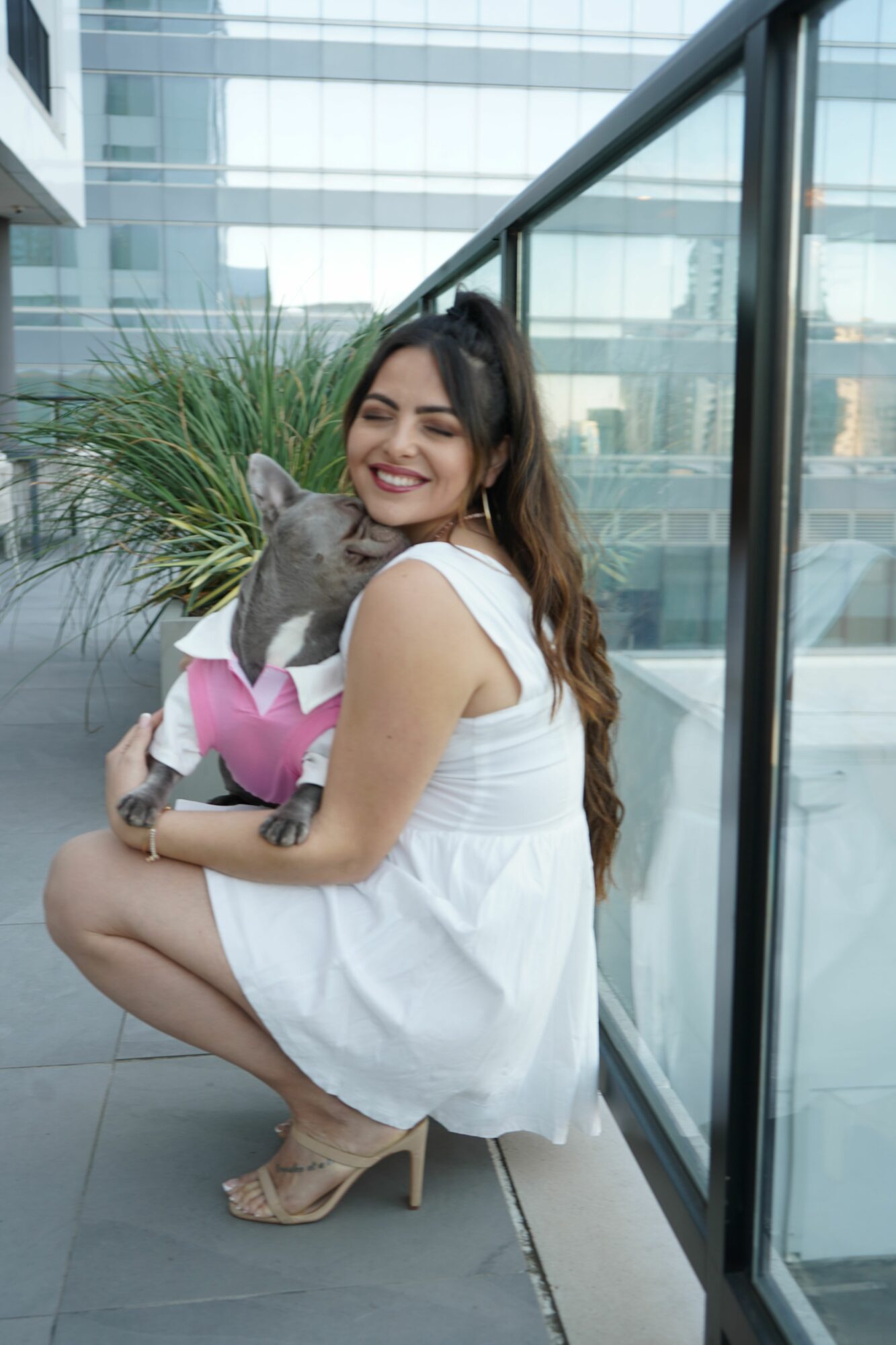 Woman in white dress sitting on floor with a gray cat, near large windows and plants, smiling at the camera.