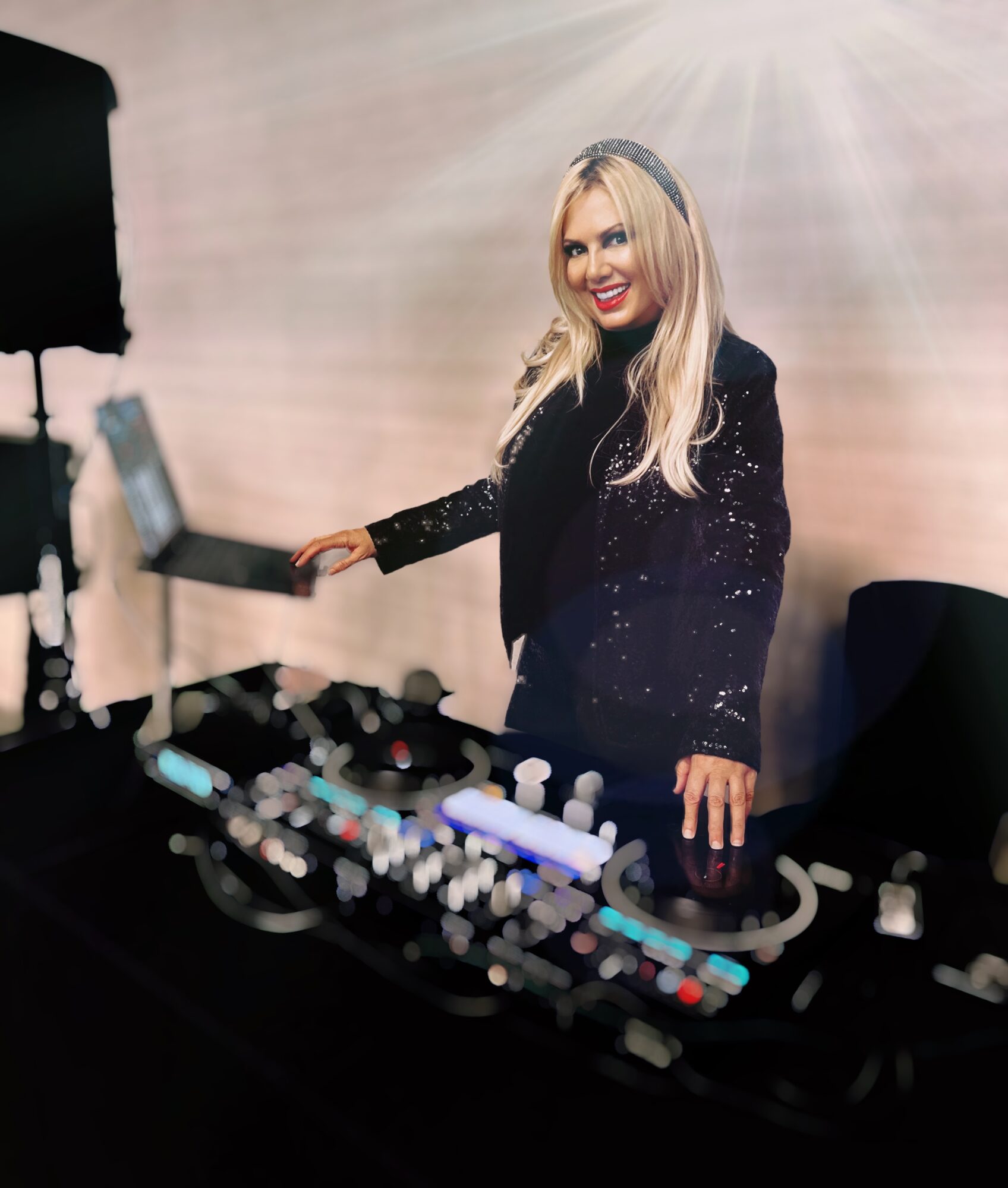 A woman with long blonde hair and a black sequined jacket stands behind DJ equipment, smiling at the camera.