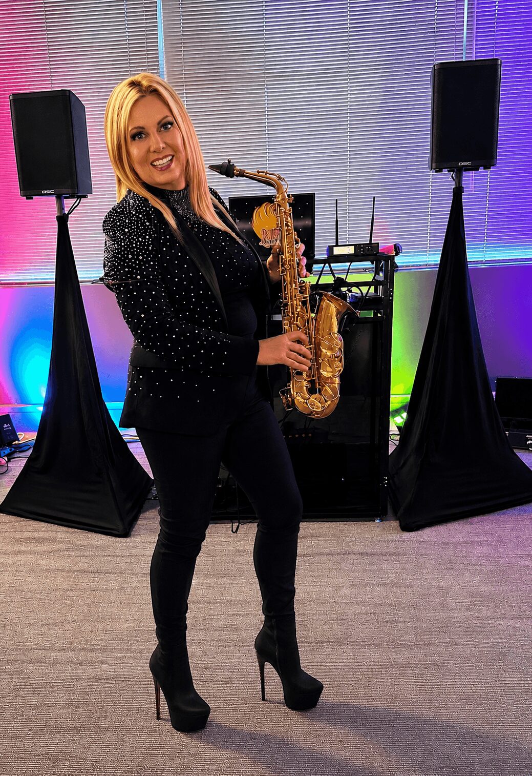 Woman with blonde hair holding a saxophone, standing between two large speakers, smiling, in a colorful room.