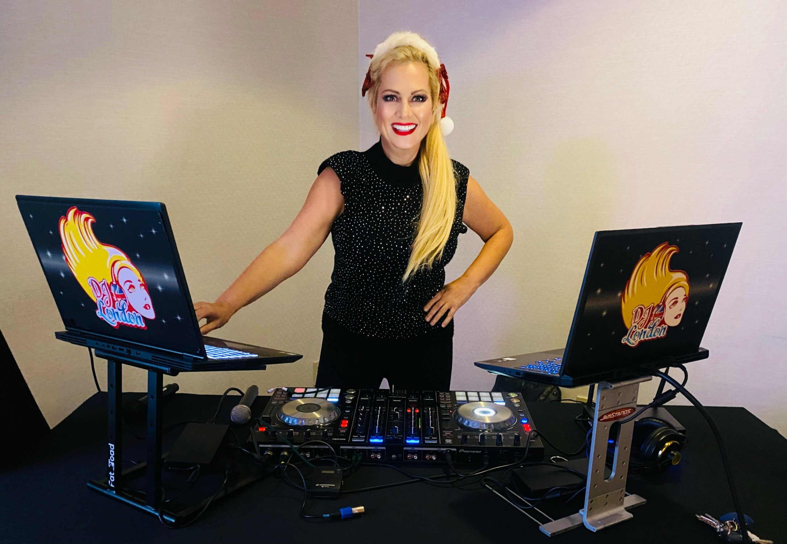 Woman DJ with two laptops featuring colorful cartoon characters, standing behind DJ equipment, smiling, with a plain background.