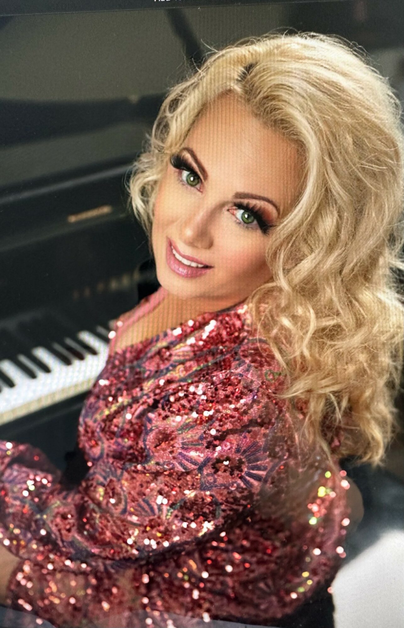 Woman with curly blonde hair smiling, wearing a sparkly red dress, sitting at a piano.