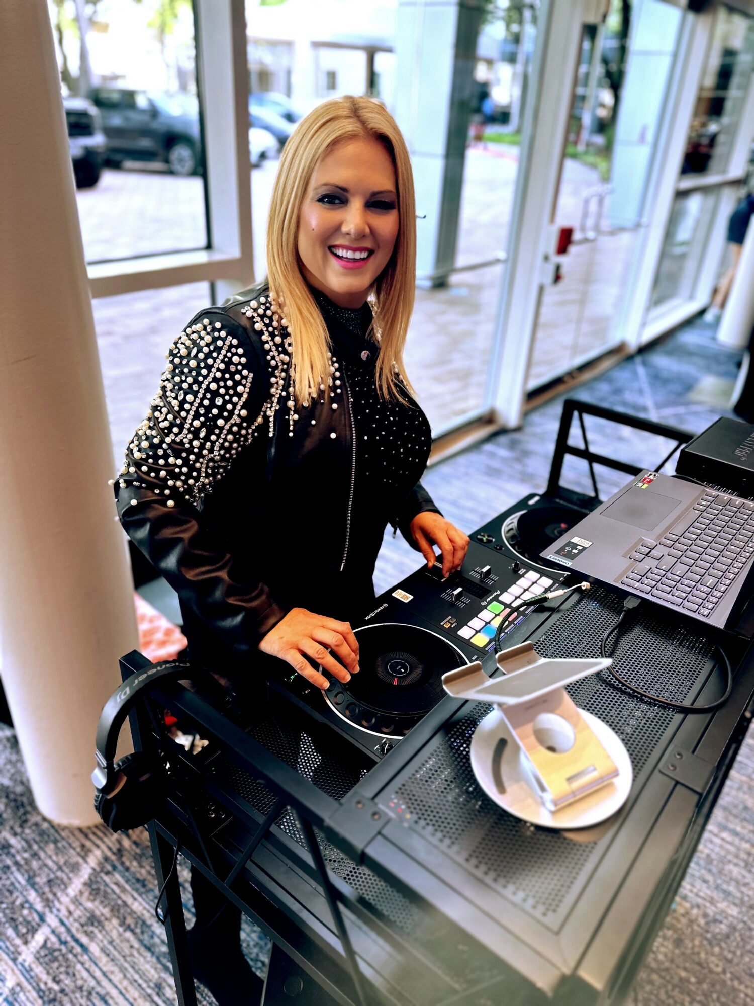 Woman with blonde hair smiling at DJ equipment in a modern indoor space with large windows.