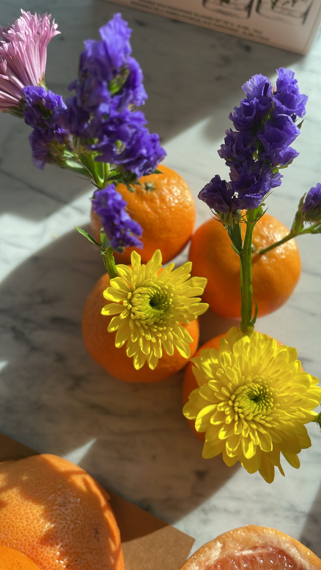 Orange fruits with purple, yellow, and pink flowers arranged on a surface, sunlight casting shadows.