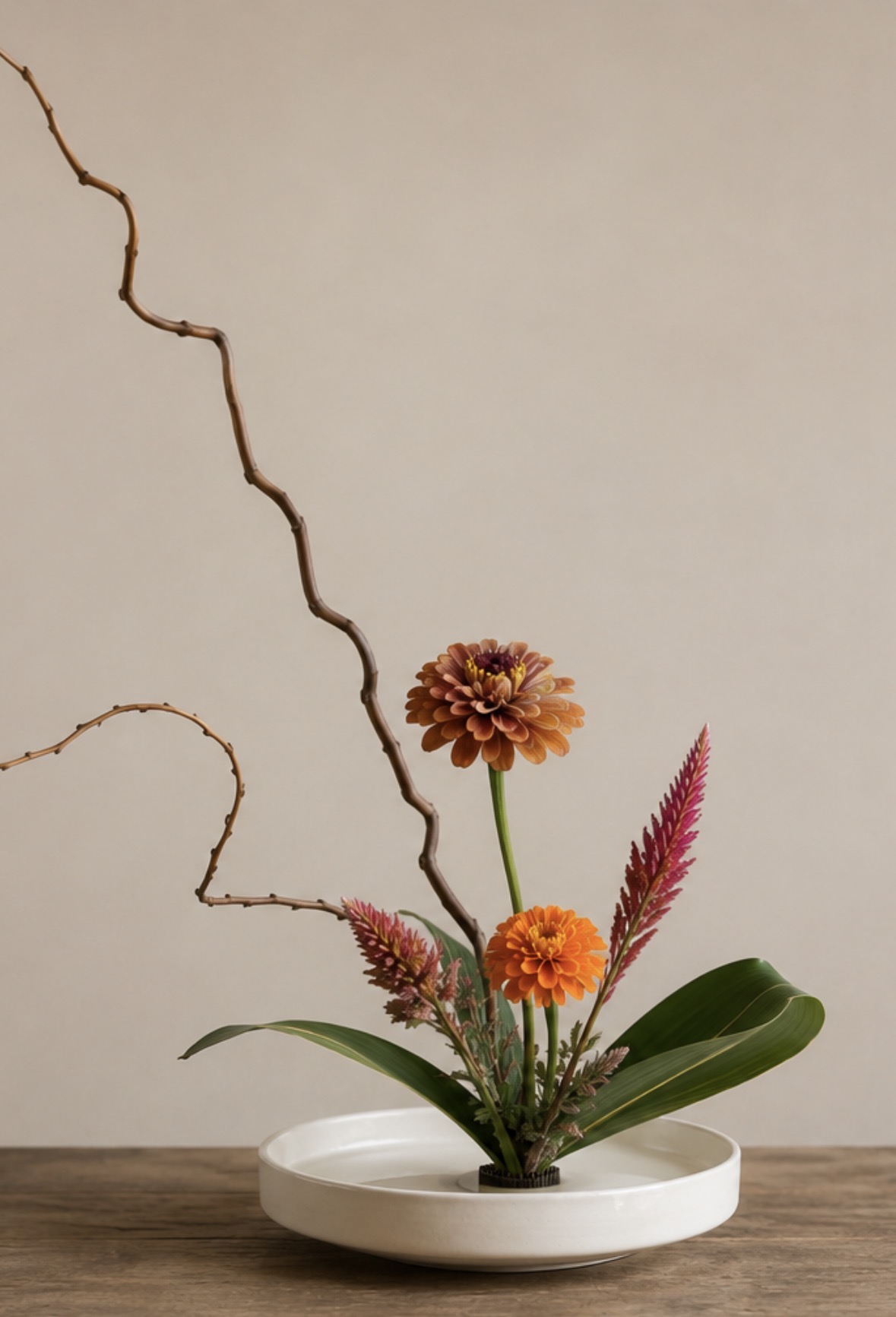 Flower arrangement with orange and pink flowers, green leaves, in a white dish on a wooden surface, against a plain wall.