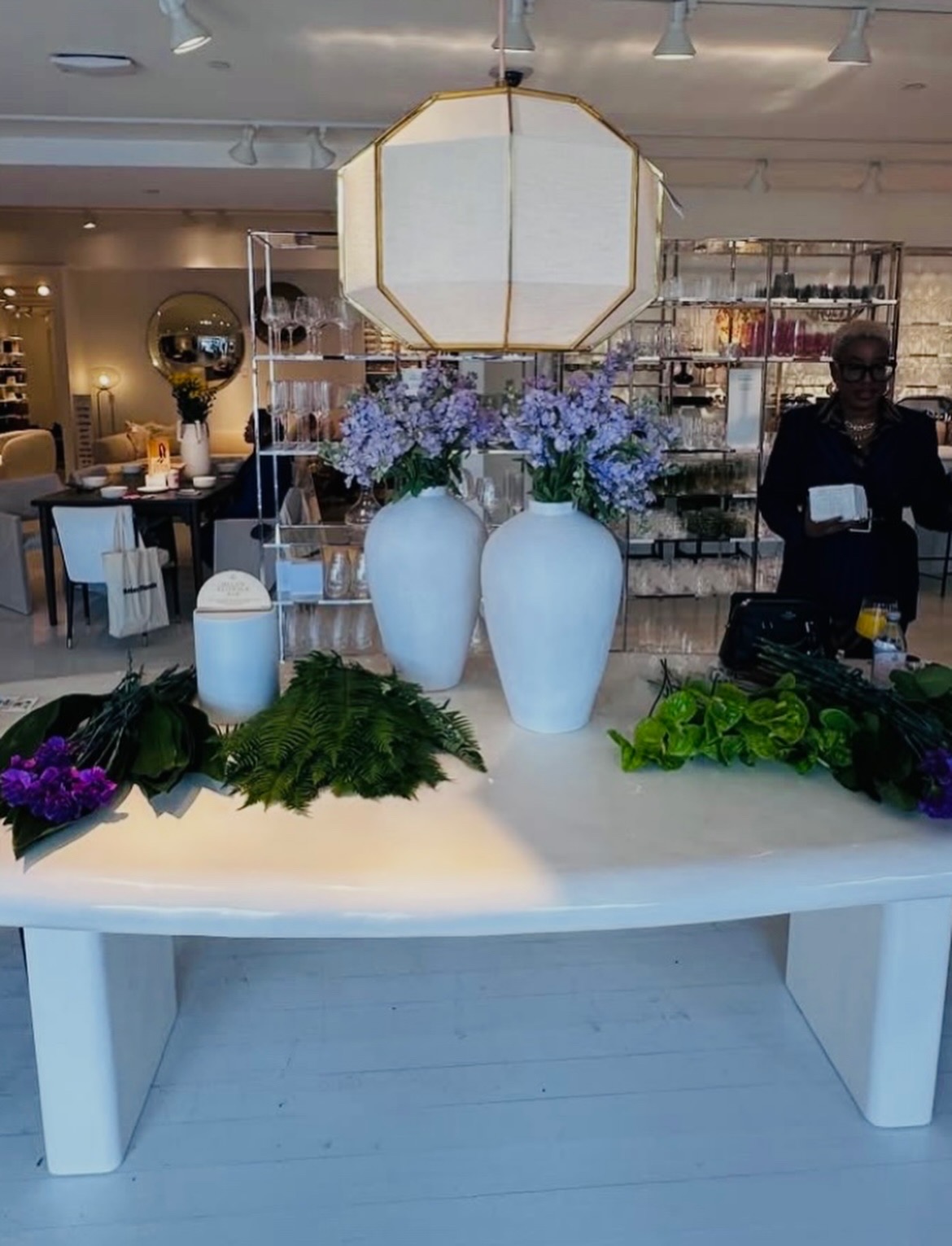 Decorative table with three white vases filled with purple and blue flowers, candles, and greenery, in a modern interior space.