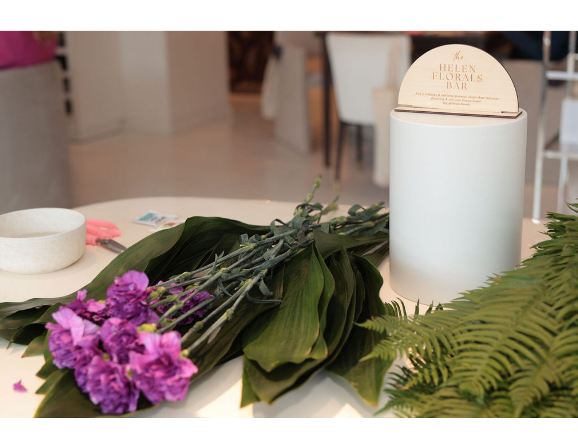 Bouquet of purple flowers and green leaves on a table with a white cylindrical container and a fern leaf, indoors.