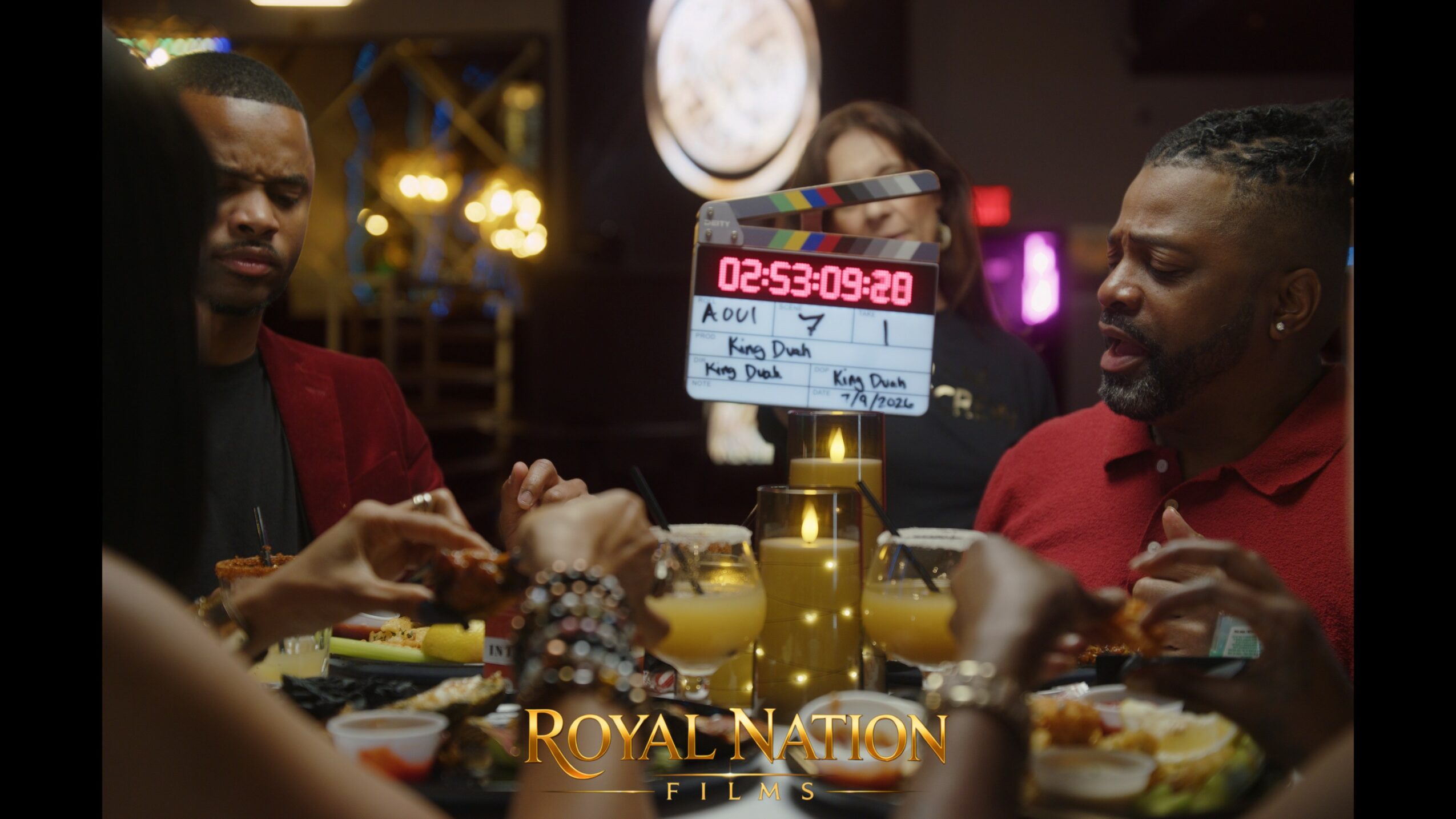 Two men sit at a table with food and drinks, a film slate is visible in the background, and a clock shows 02:53:09.