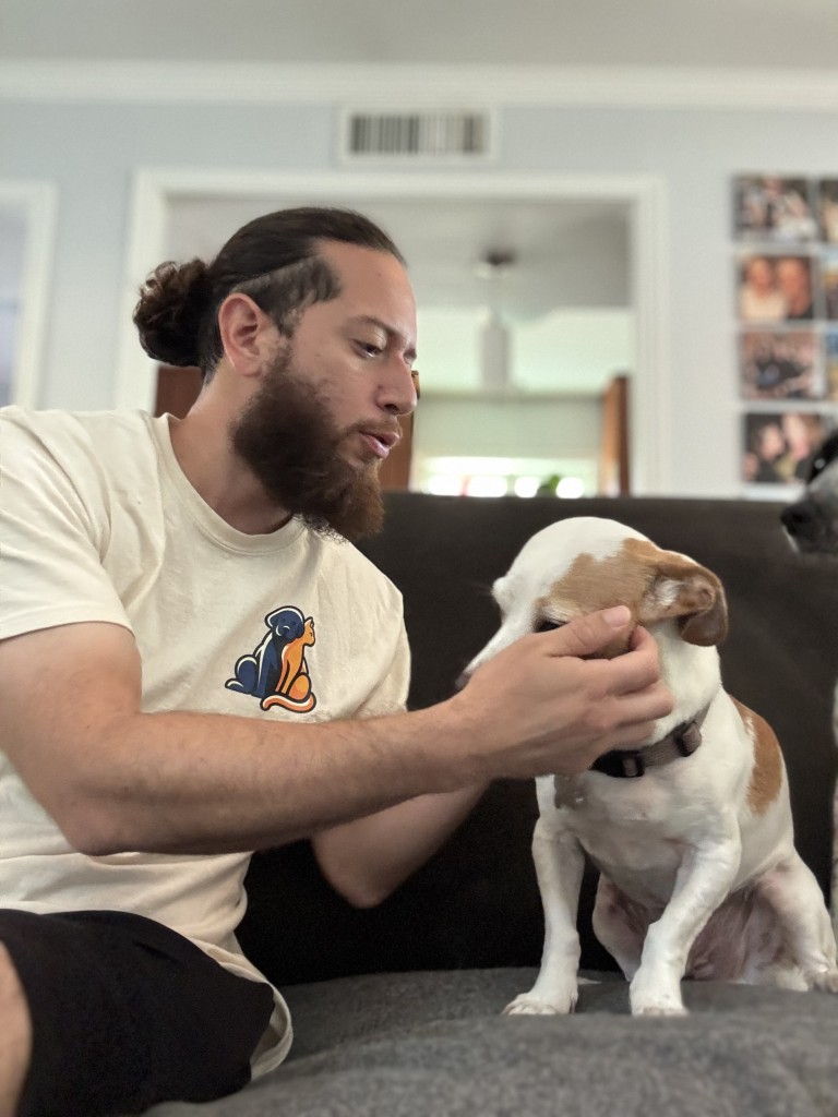 Man with a beard and bun petting a small dog on a couch in a living room.