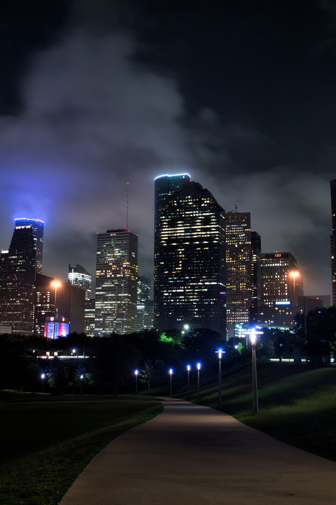 Nighttime city skyline with illuminated buildings and cloudy sky, viewed from a park with a pathway and streetlights.