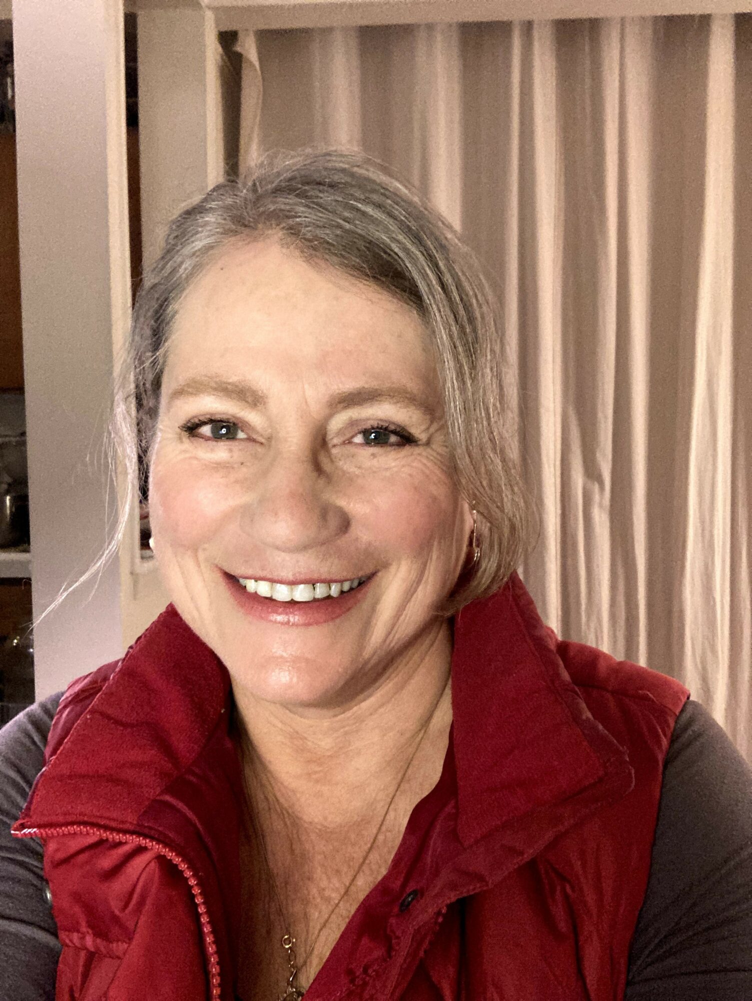 Smiling woman with gray hair in a red vest, indoors with curtains in the background.