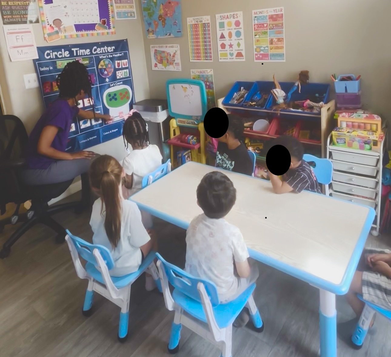 Children sit around a white table in a classroom with educational posters and toys on the walls.