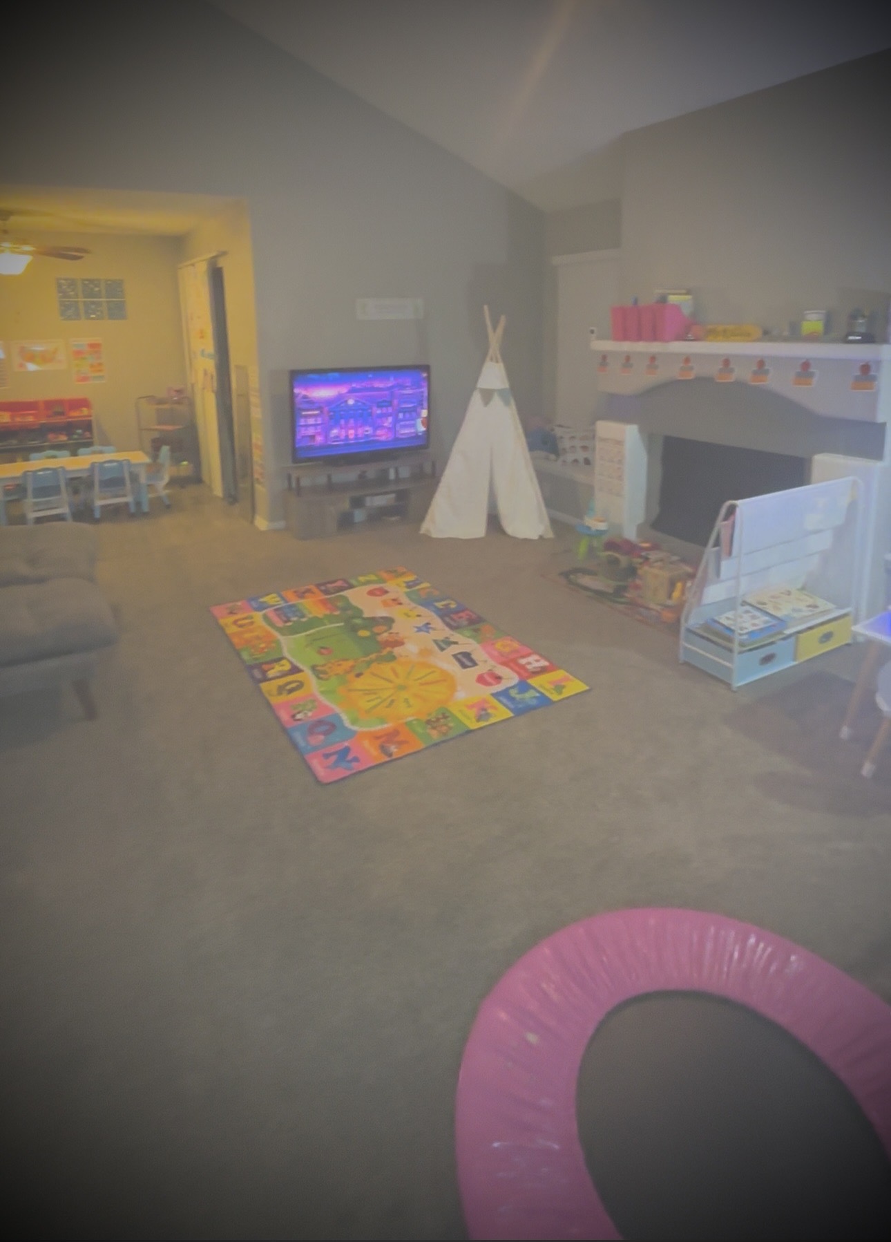Living room with colorful rug, TV, teepee, and children's toys, viewed from the front. Bright yellow room visible through doorway.