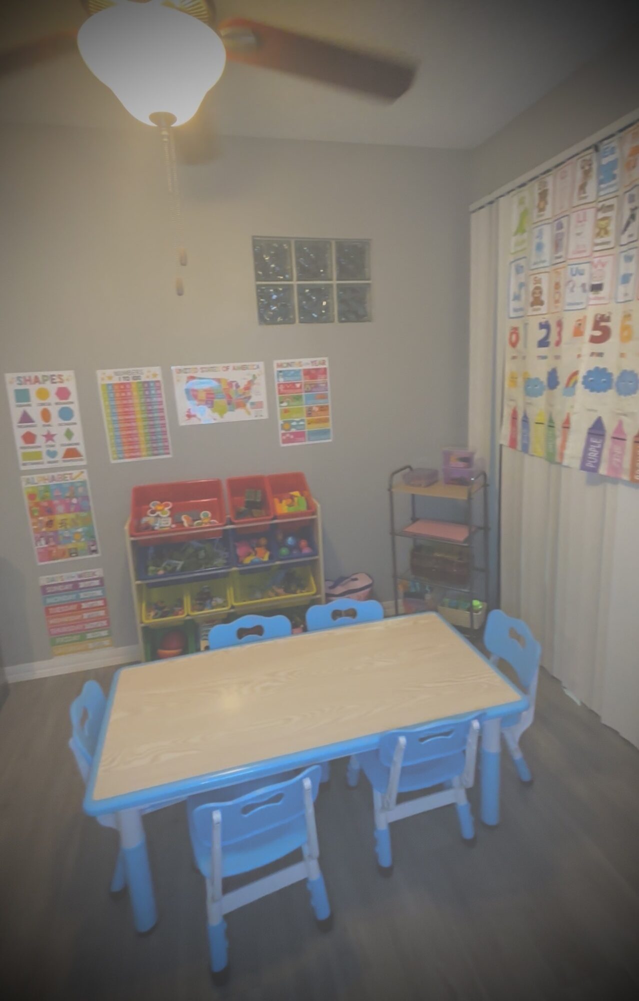 Classroom with a table, six blue chairs, educational posters, and a shelf with toys and books, illuminated by a ceiling light.