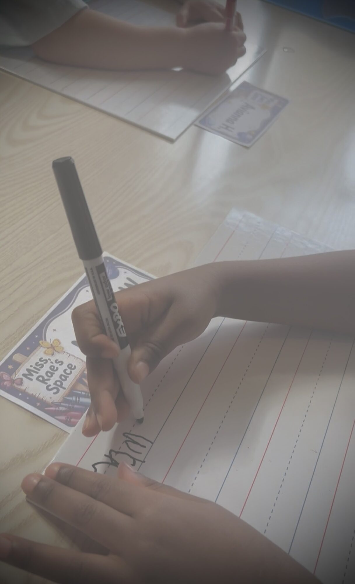 Child's hand writing on lined paper with a marker, with a card and paper on a light-colored table.