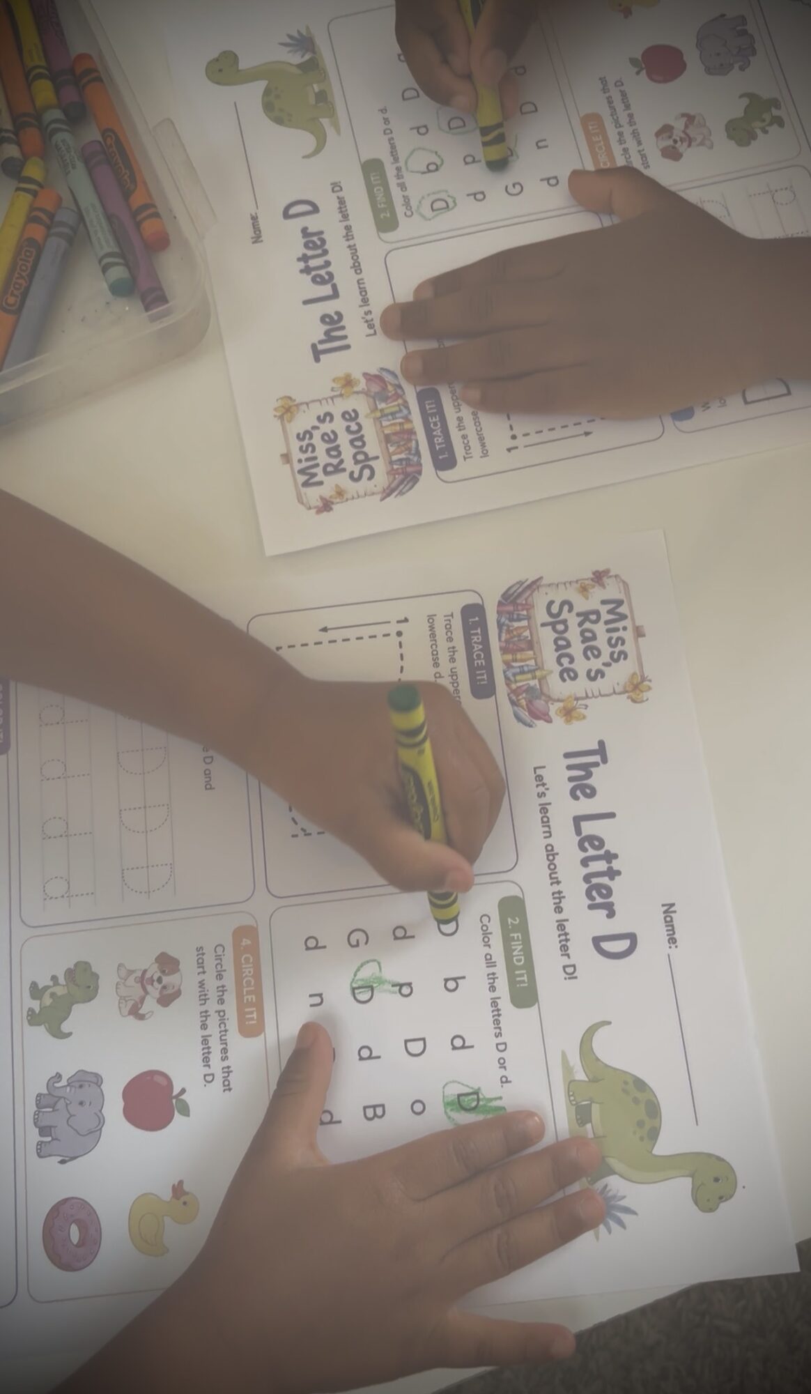 Two children work on a letter D worksheet with colorful illustrations and writing activities, hands visible on the paper.