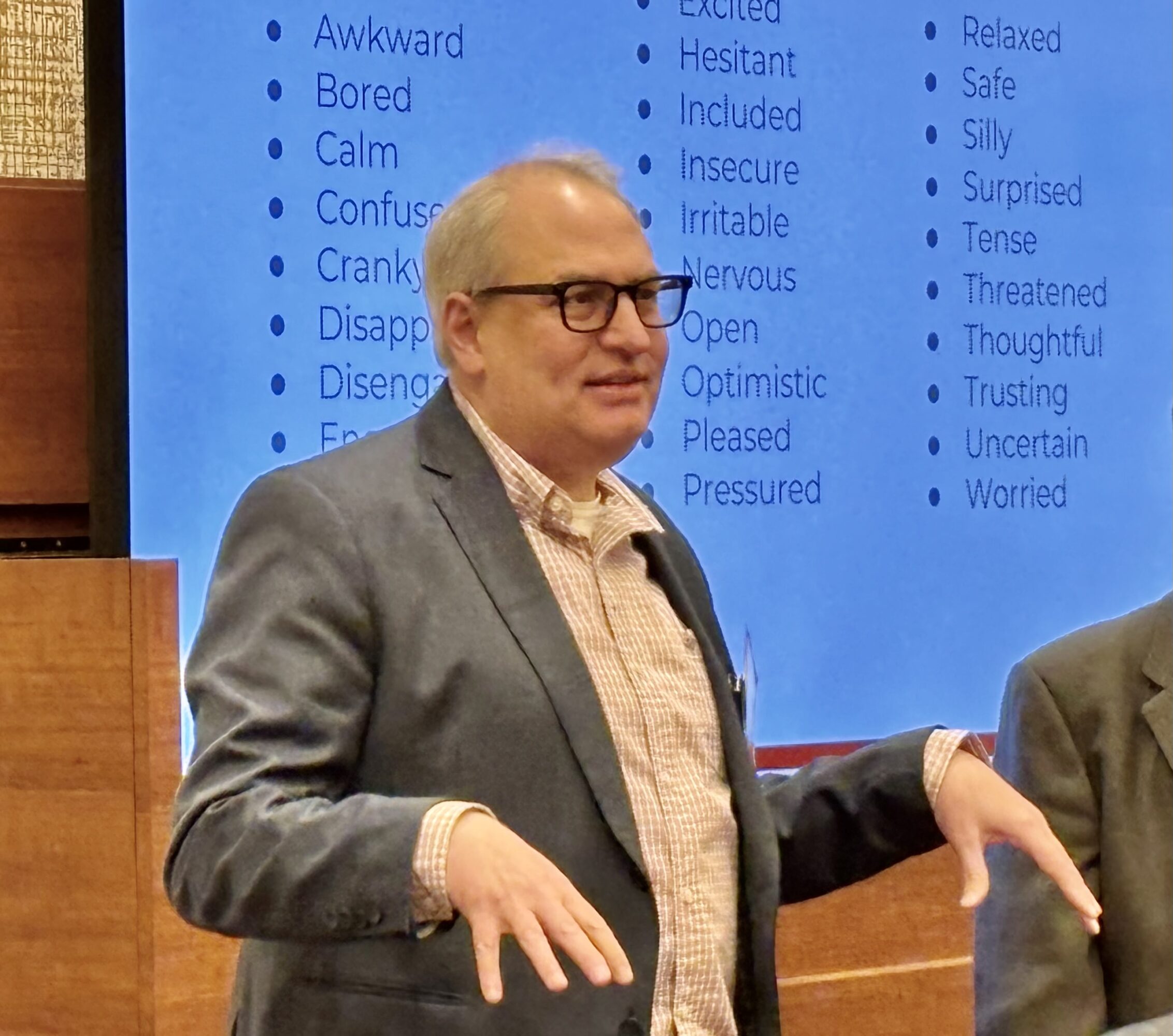 Man with glasses in a suit standing in front of a blue screen with words, gesturing with hands.