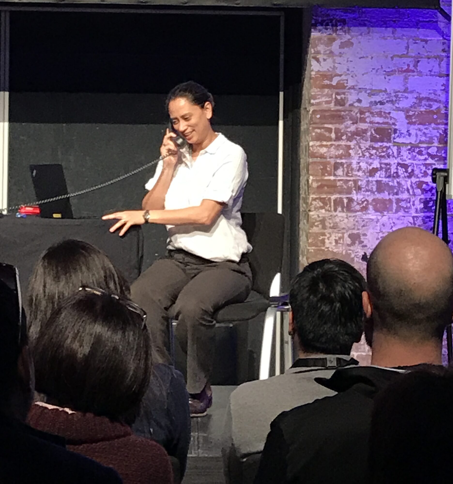 Woman sitting on a stool, talking on a phone, with an audience in front, brick wall background, stage setting.