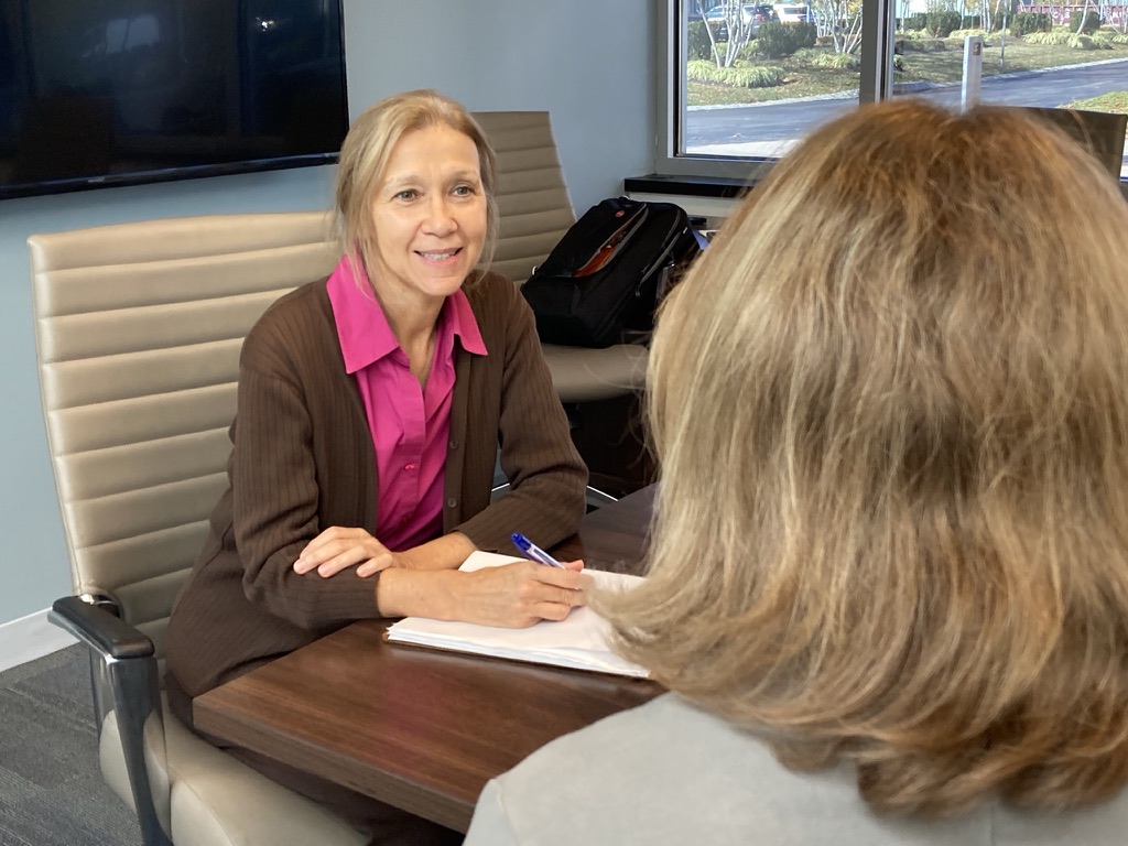 Two women sitting at a conference table, one smiling and holding a pen, in a modern office setting.