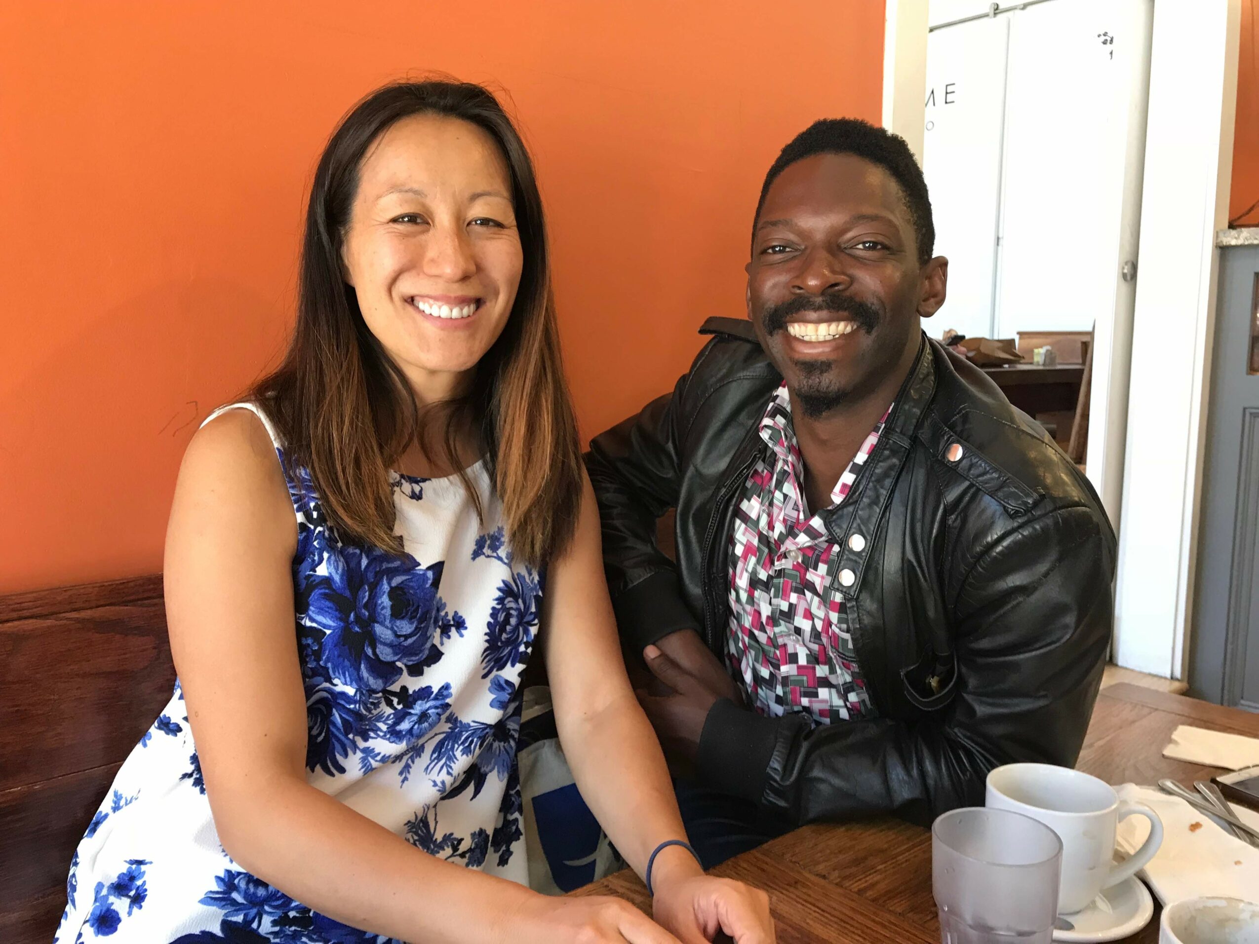 Two people smiling, sitting at a table with cups, in a room with orange and white walls.