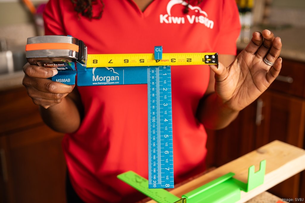 Person in red shirt holding a blue and yellow measuring device, measuring height, with a green object on a wooden surface.