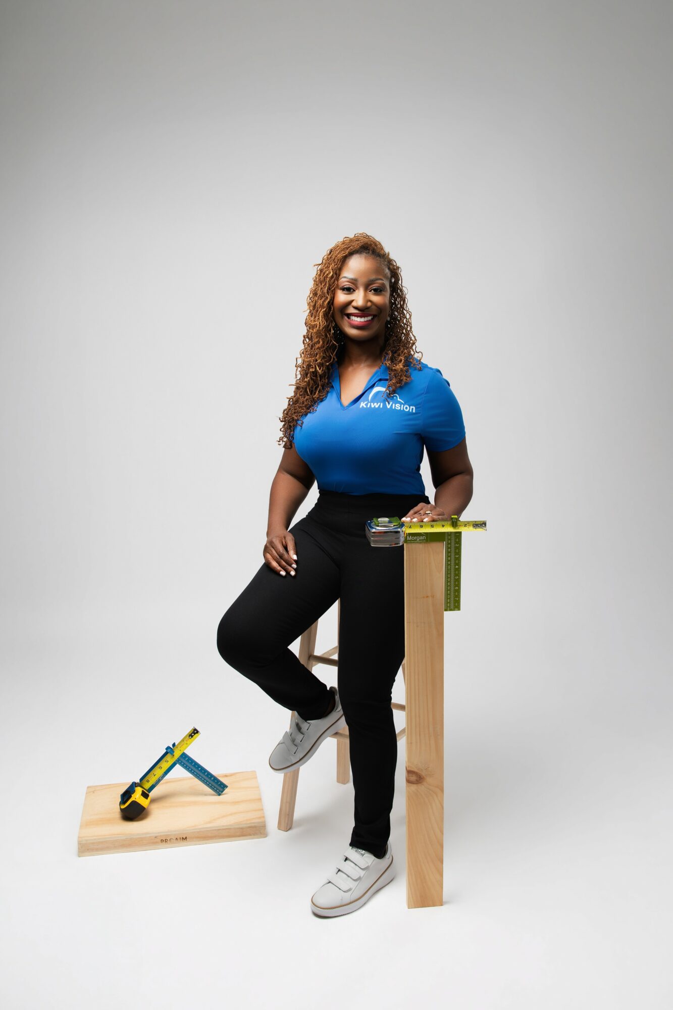 Smiling woman in blue shirt standing with one foot on a small wooden platform, surrounded by tools, against a plain background.