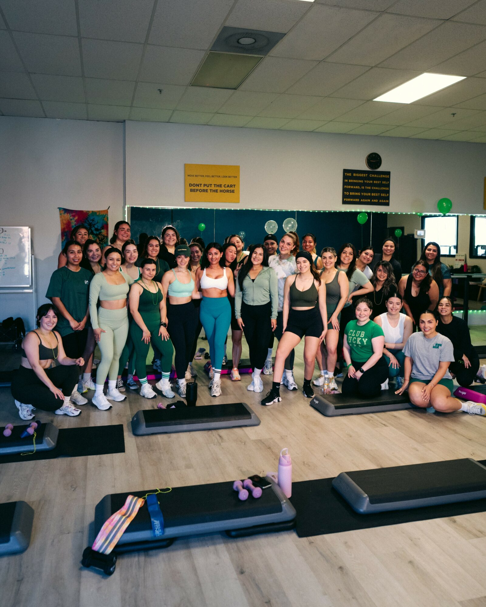 Group of people in workout attire standing in a fitness studio, some on exercise mats, with equipment and mirrors in background.