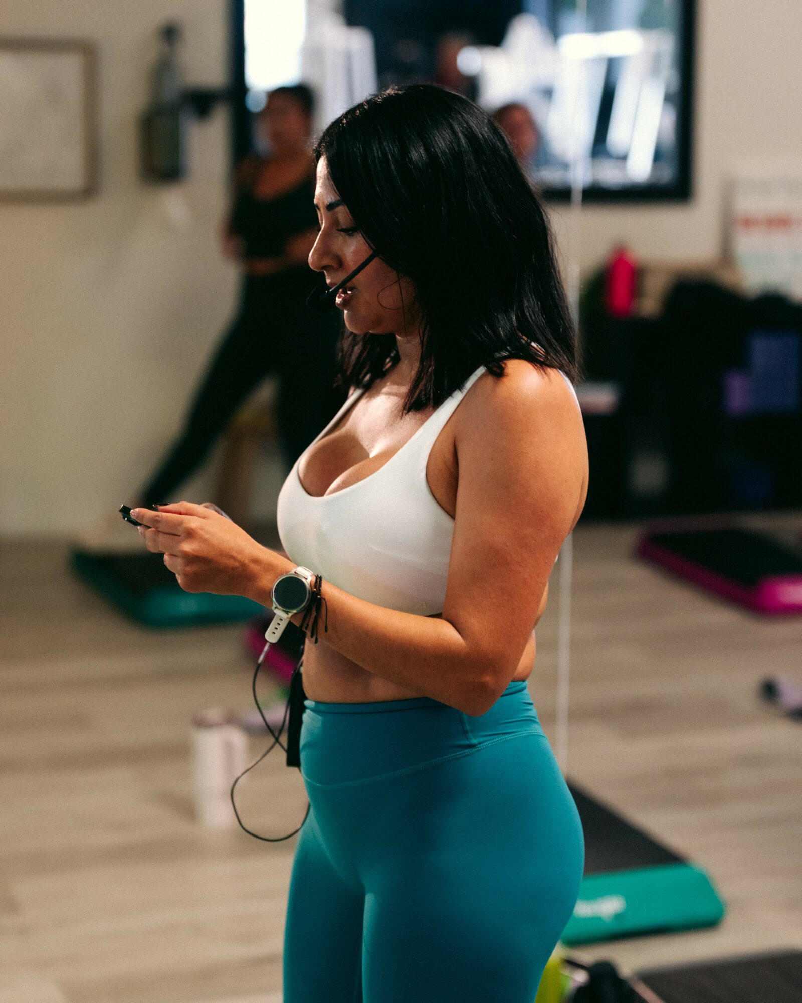 Woman with black hair in a white top and blue leggings looking at her phone in a gym.