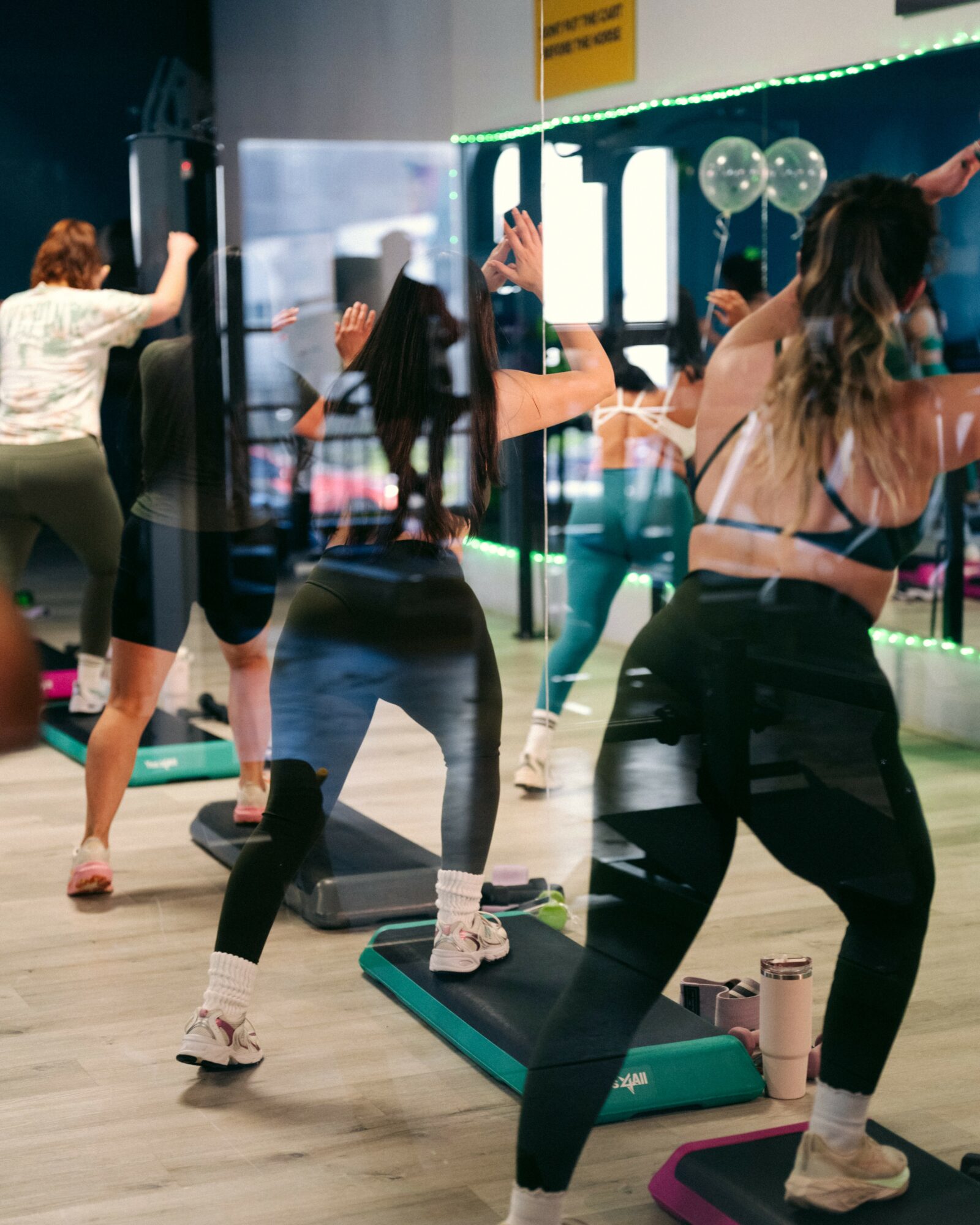 Group of people exercising on step platforms in a fitness class, with mirrors and balloons in the background.