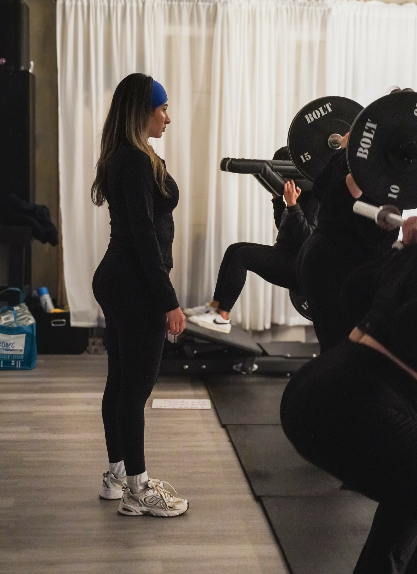 Woman standing in front of a person lying on a bench, lifting weights in a gym, with a curtain background.