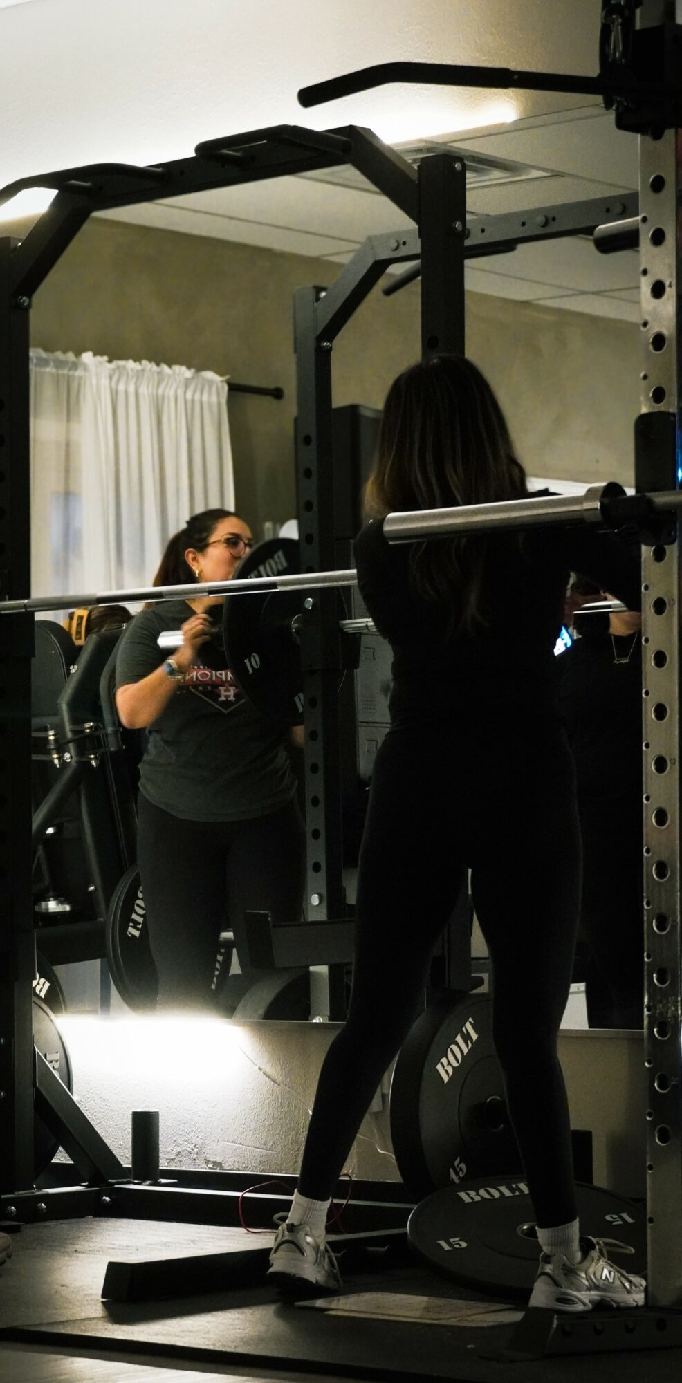 Two women in a gym, one using a weight machine and the other standing nearby, with a mirror reflecting the scene.
