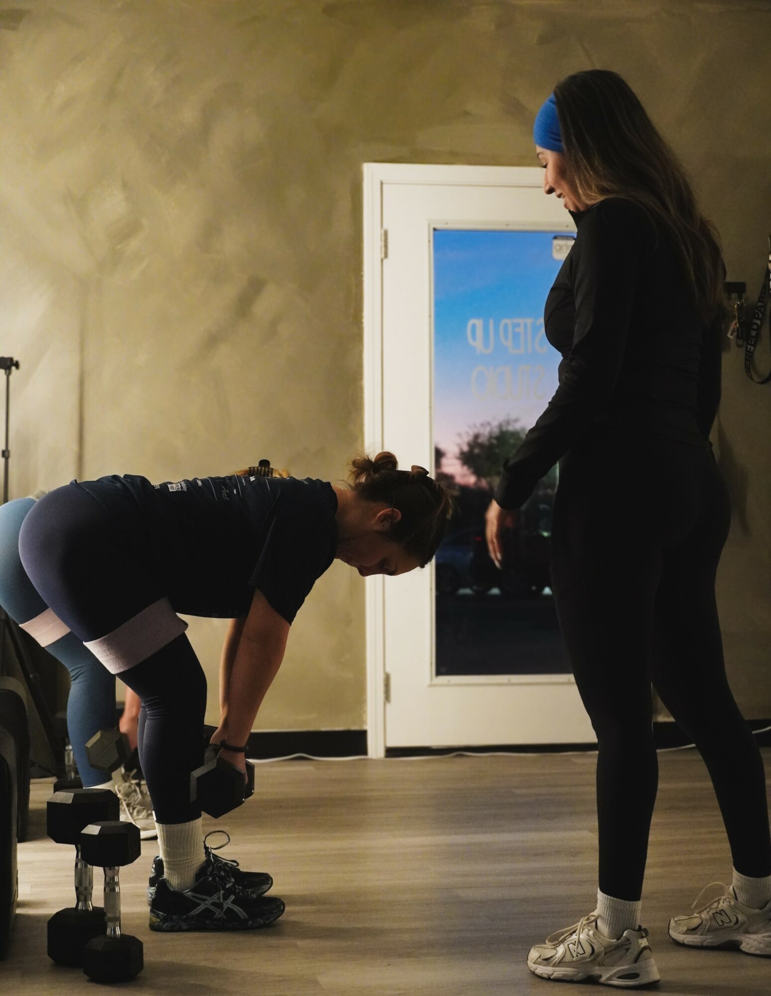 Two women in workout clothes in a gym, one bending forward, the other standing nearby, with a door and window in the background.