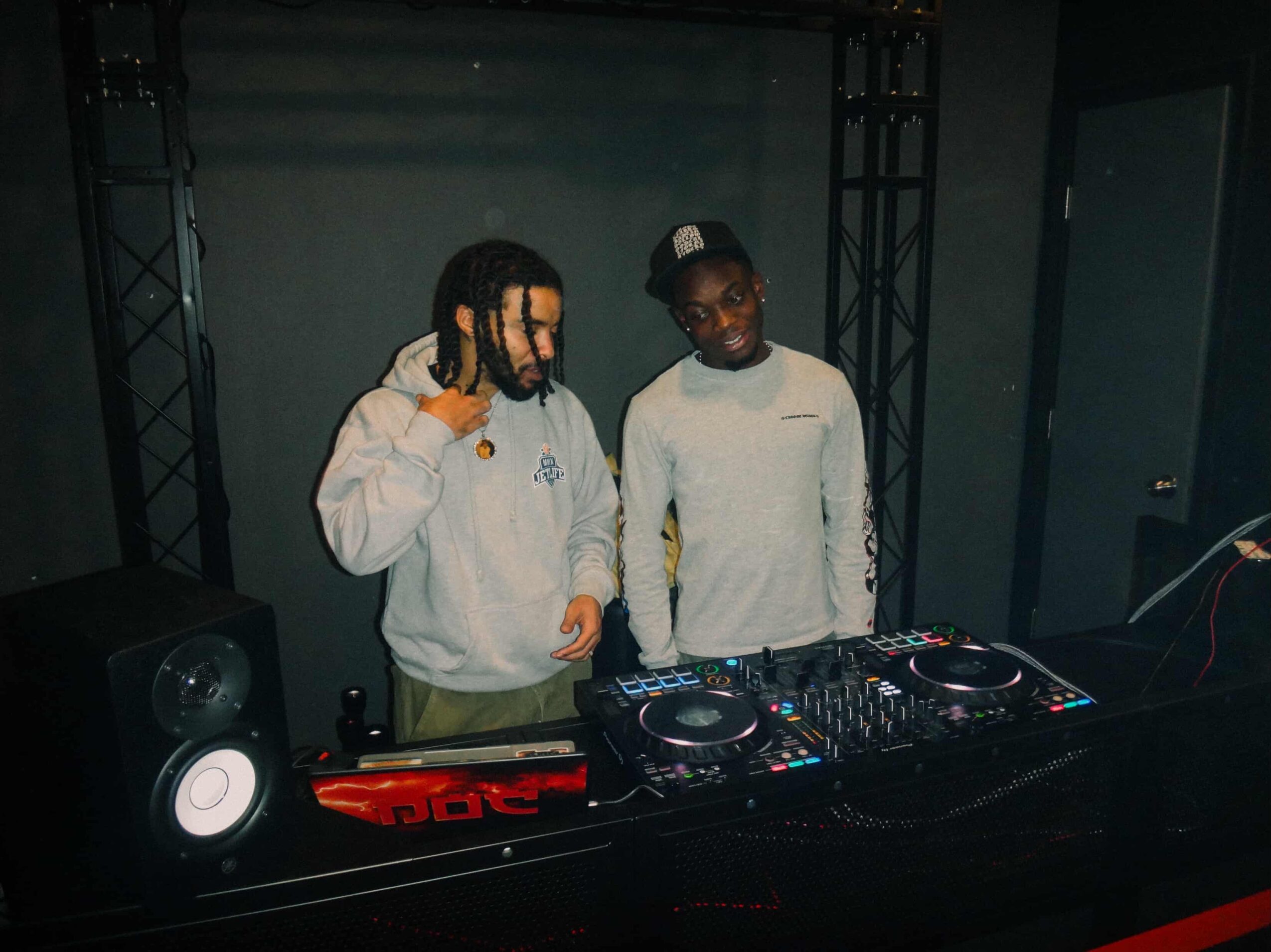 Two people stand behind DJ equipment, with speakers and a dark background.