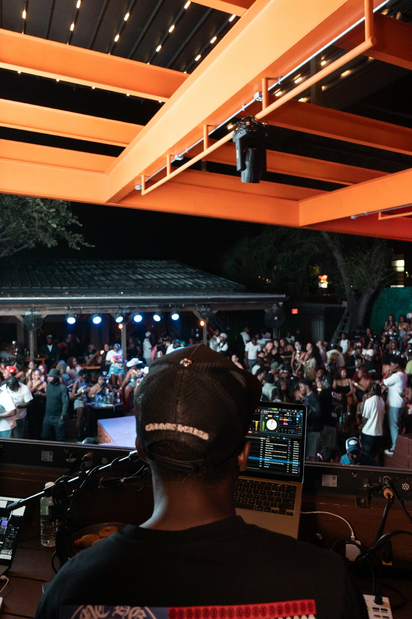 DJ performing at an outdoor event with a crowd, stage, and orange overhead structure, nighttime setting.