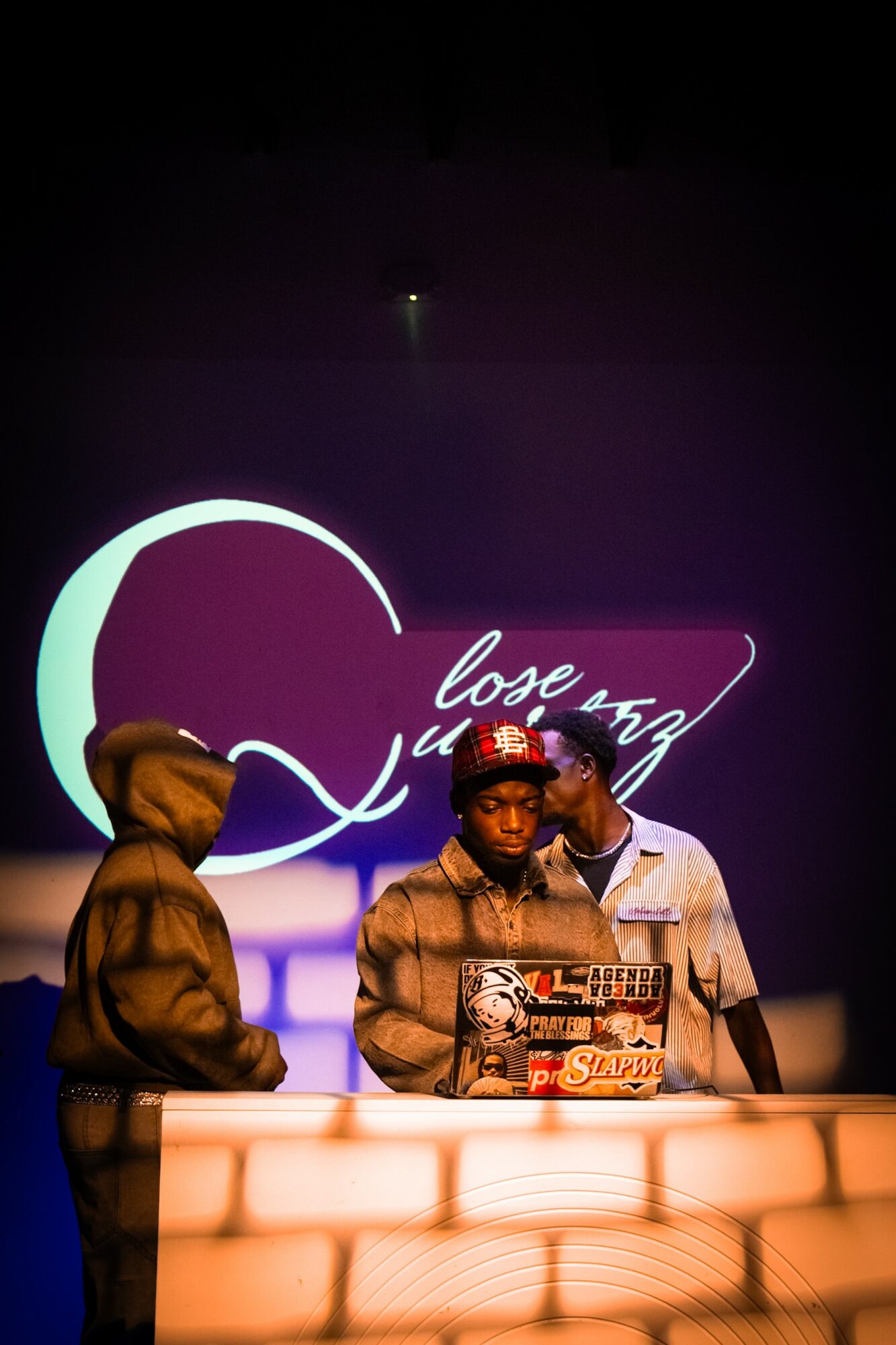 Three people stand behind a lit-up counter with a dark background and neon sign reading 'Love Song'.