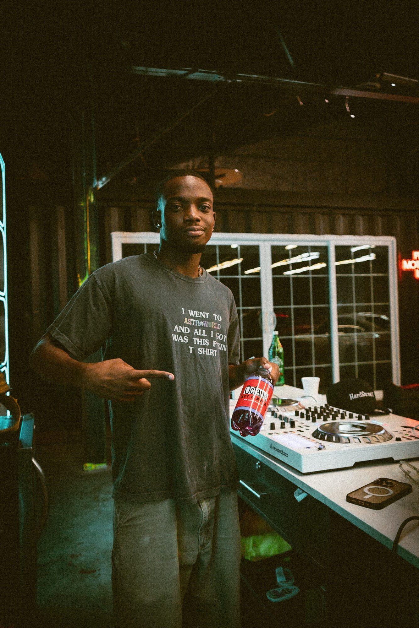 Young man standing next to DJ equipment in a dimly lit room, pointing at a bottle on the table.