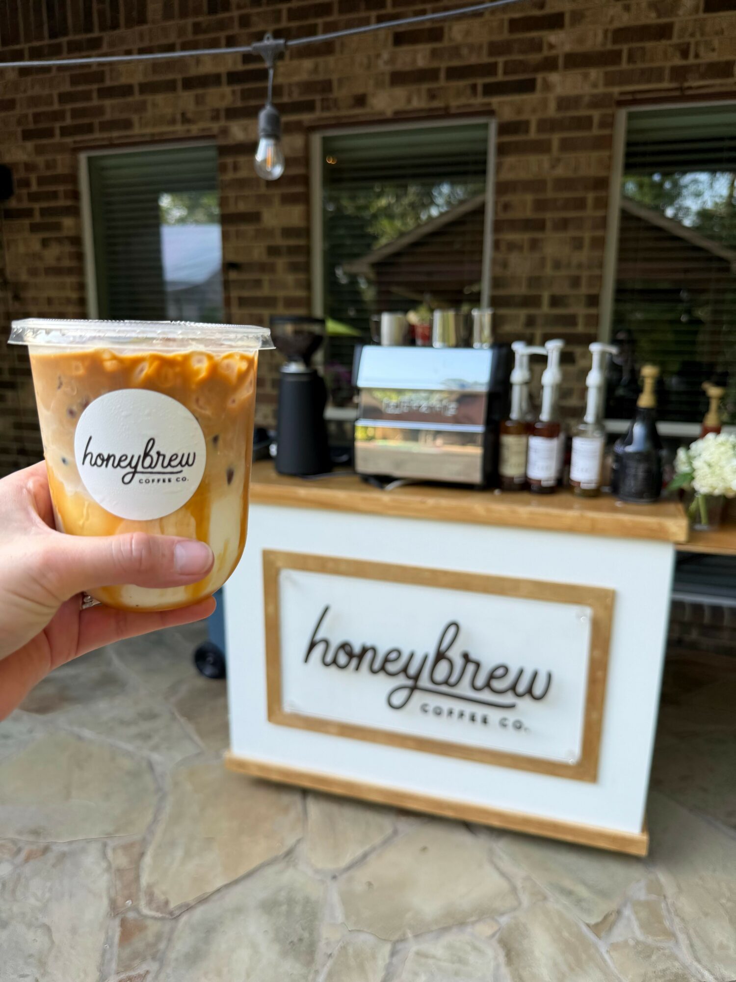Person holding a cup of coffee in front of a sign with the word 'homebrew' on it, outside a building with brick walls.