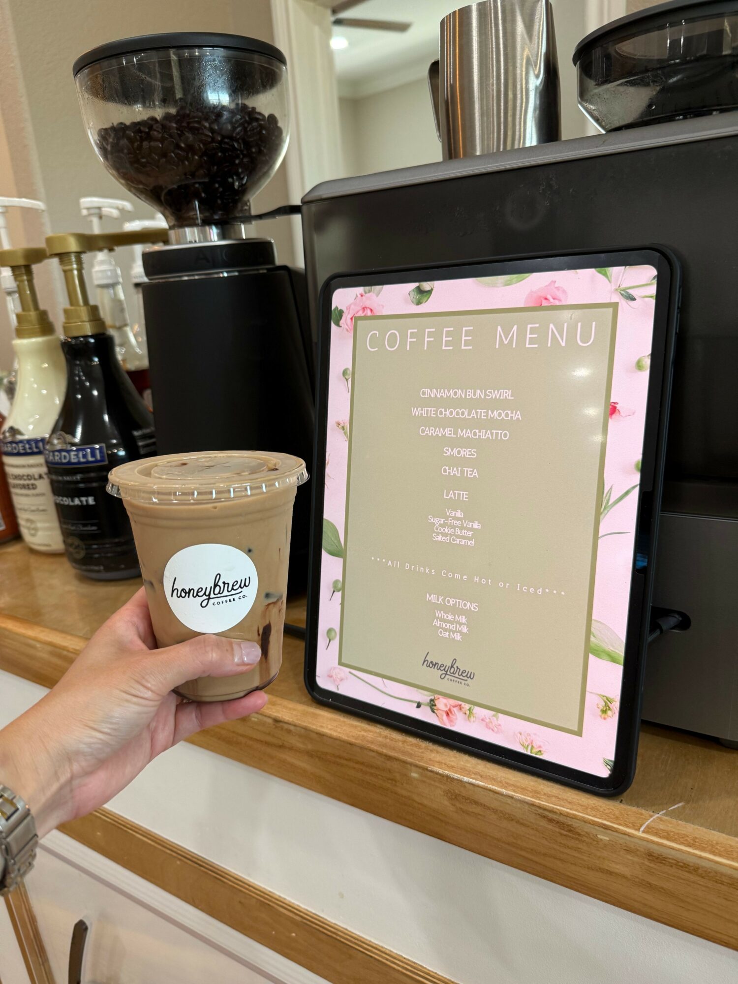 Person holding a cup of coffee in front of a digital menu board at a cafe, with a black tumbler nearby.
