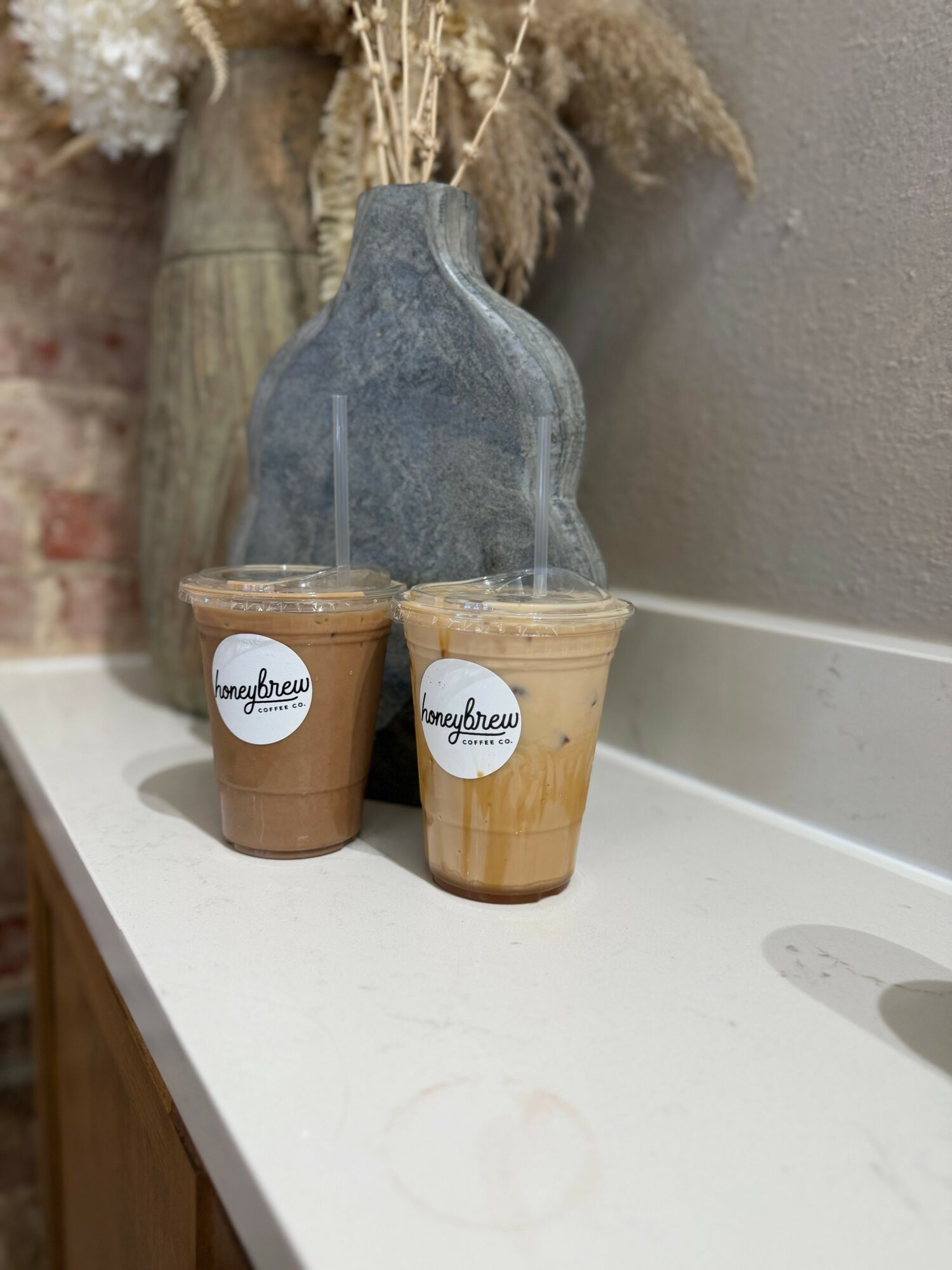 Two iced coffee drinks with straws on a white surface, behind a gray vase with dried plants, against a wall.
