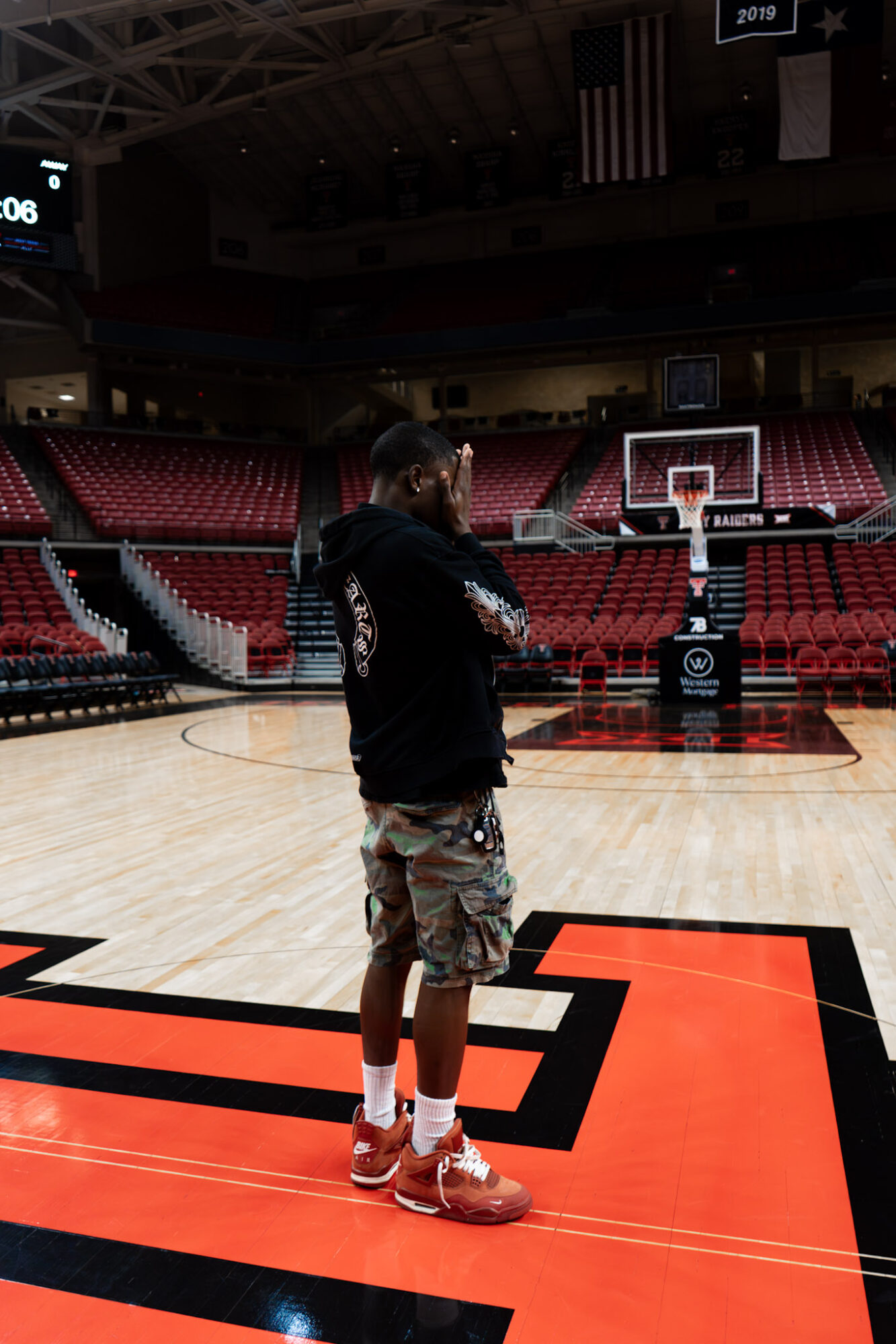 Person standing on basketball court with arms crossed, in an empty arena with red seats and a basketball hoop.