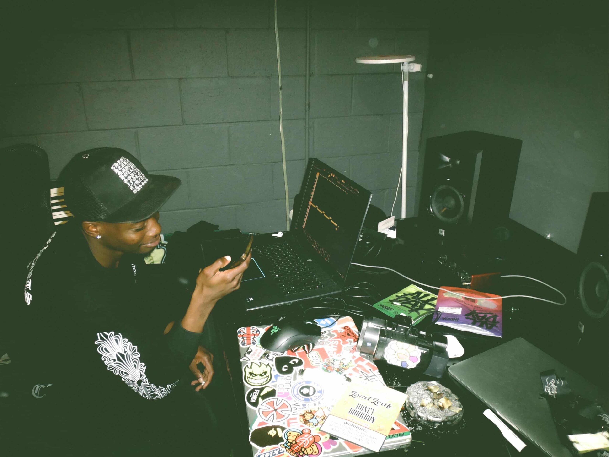 Person wearing a cap and headphones, sitting at a desk with a laptop, speakers, and various items, in a room with green walls.