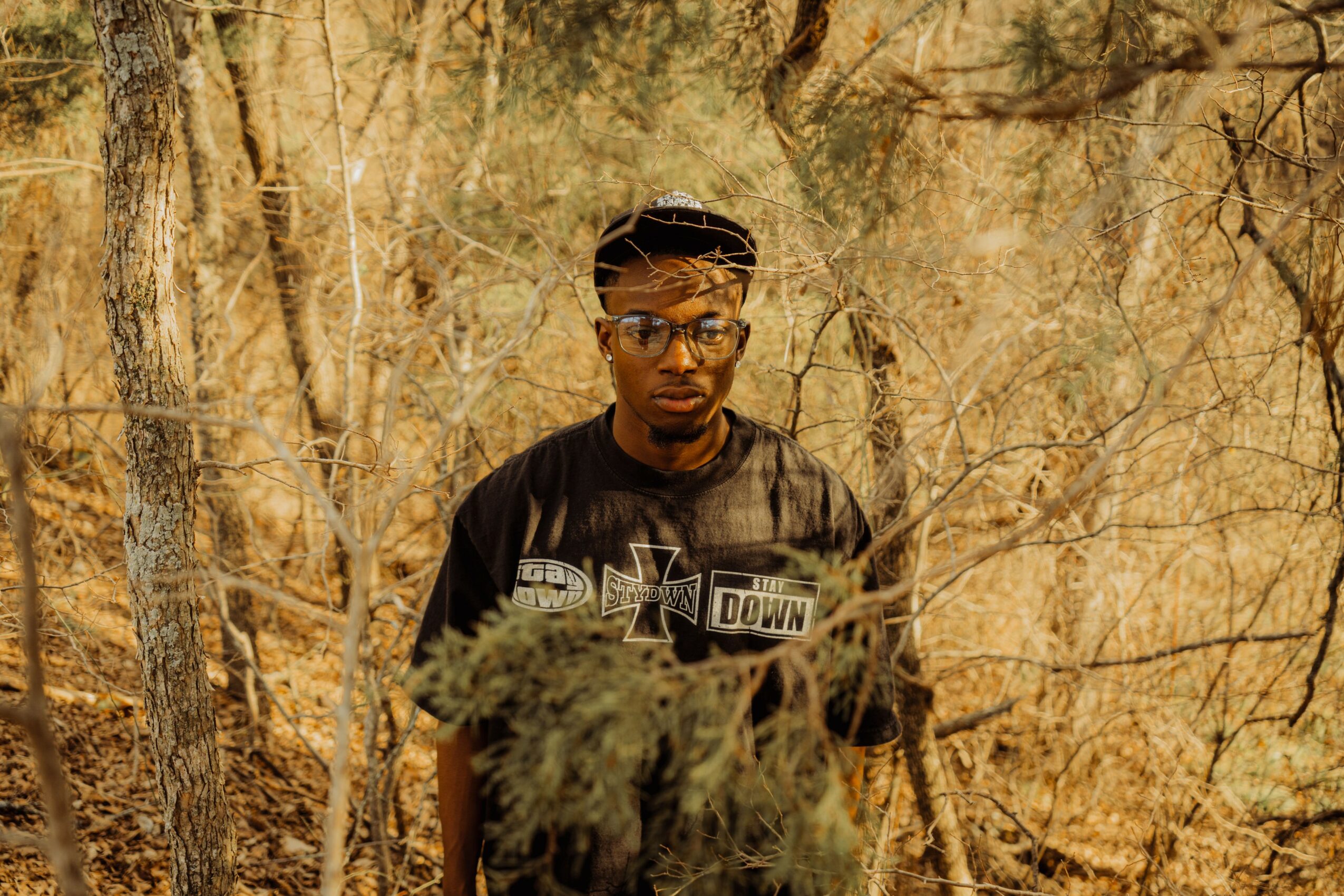Young man with glasses and a black jacket standing outdoors among trees and dry foliage.