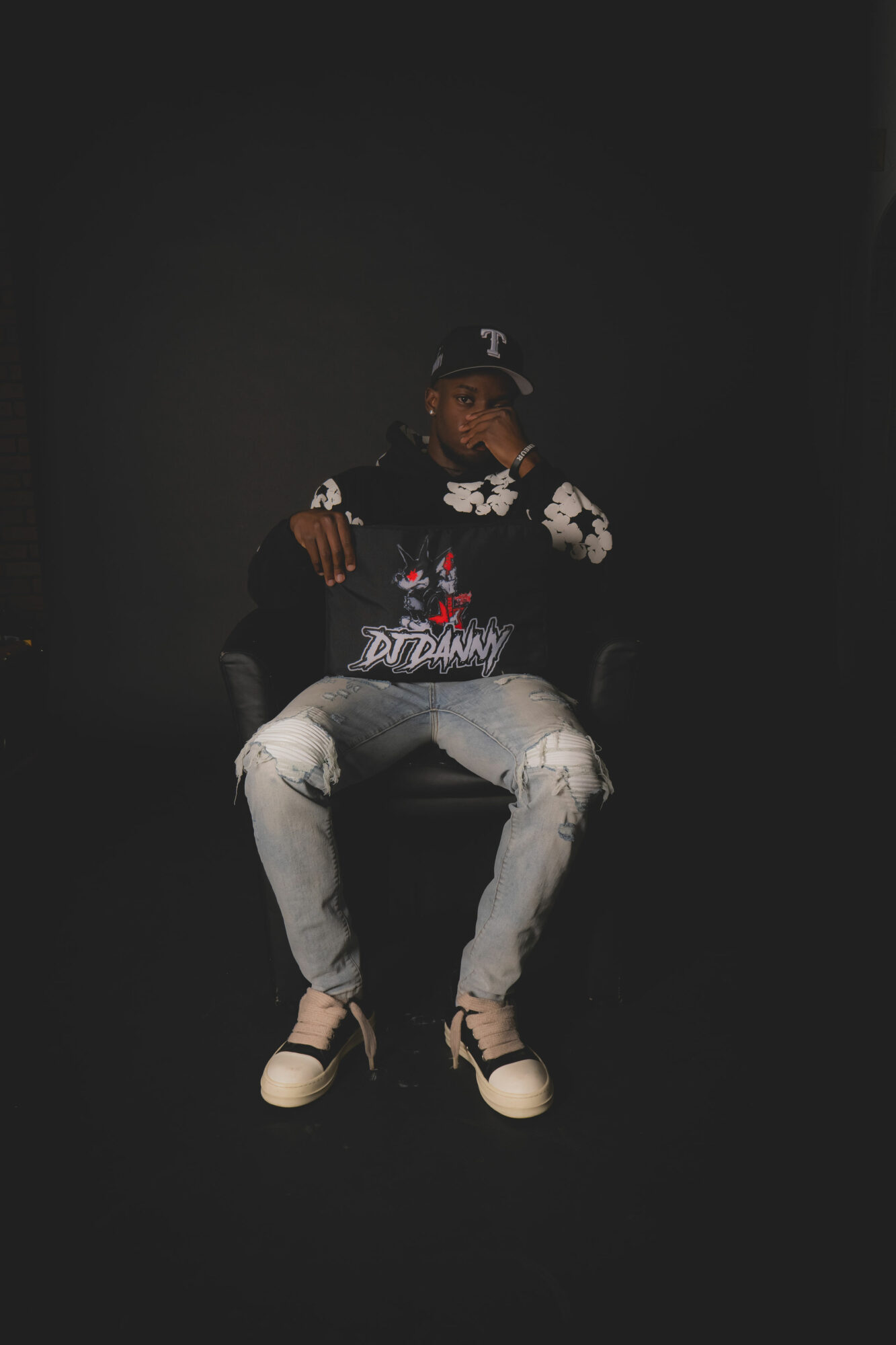 Person sitting on a chair against a dark background, wearing a cap, floral shirt, and light jeans, with sneakers.