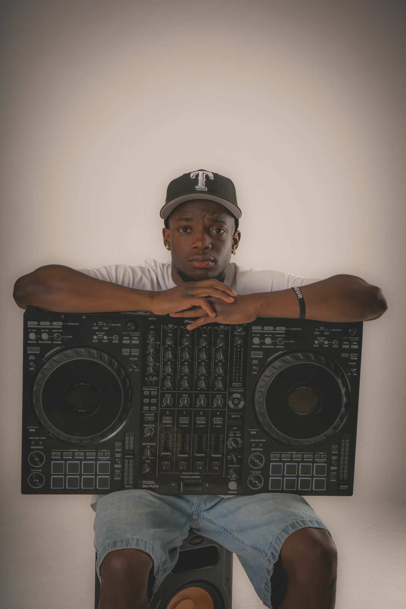 Young person wearing a cap and gray shirt, sitting behind DJ equipment, with arms resting on it, against a plain background.