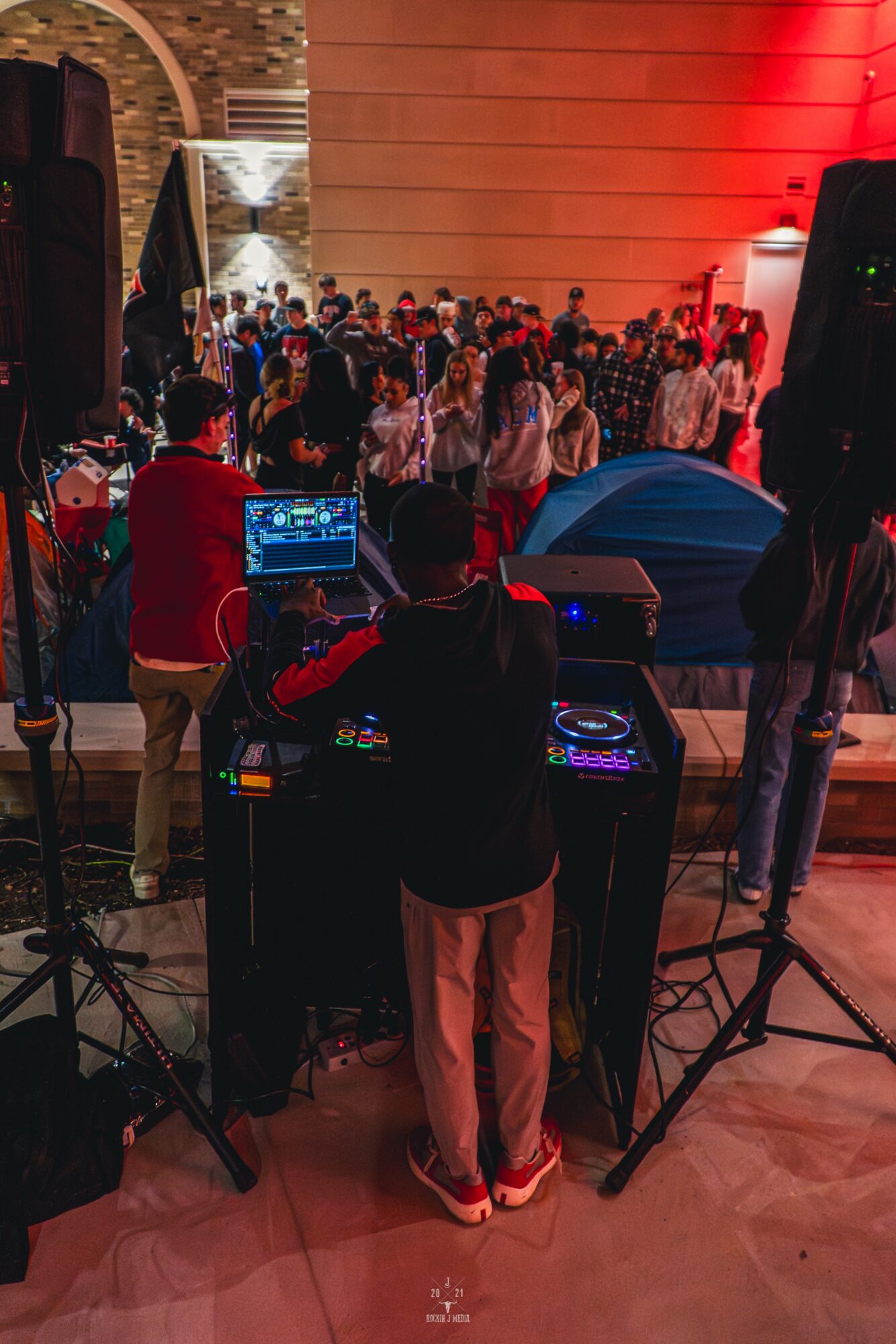DJ performing at a crowded indoor event with people dancing and socializing, warm lighting, and a brick wall in the background.