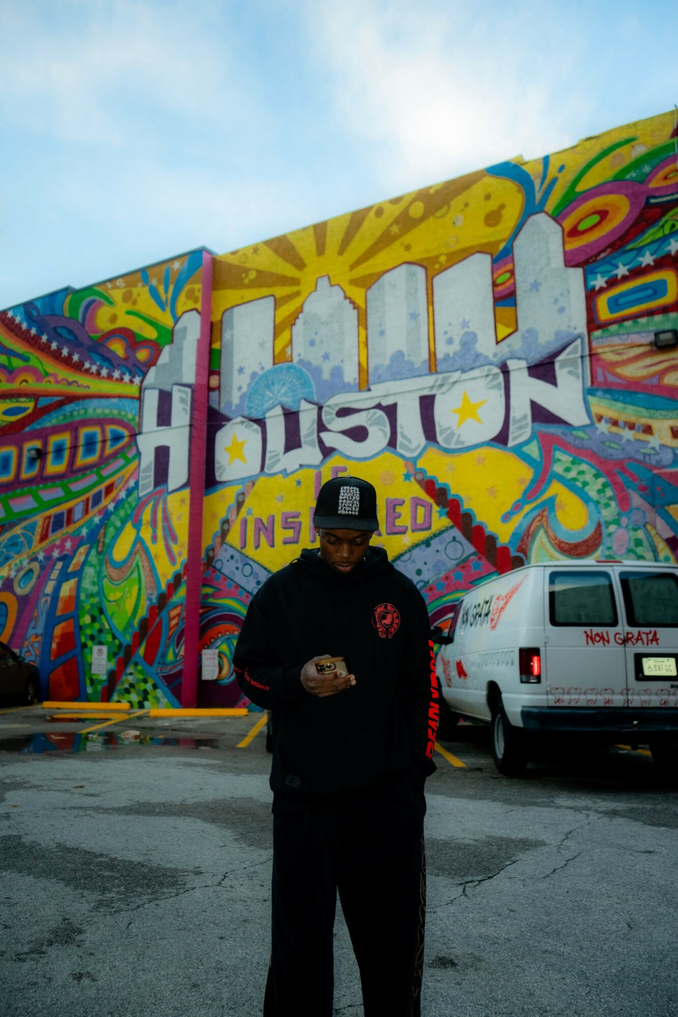 Person standing in front of a colorful graffiti mural with the word 'HOUSTON' and a white vehicle nearby.