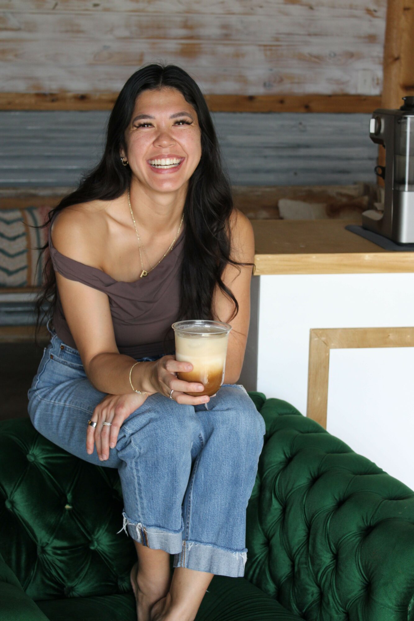 Woman with long dark hair smiling, holding a drink, sitting on a green velvet couch in a casual setting.