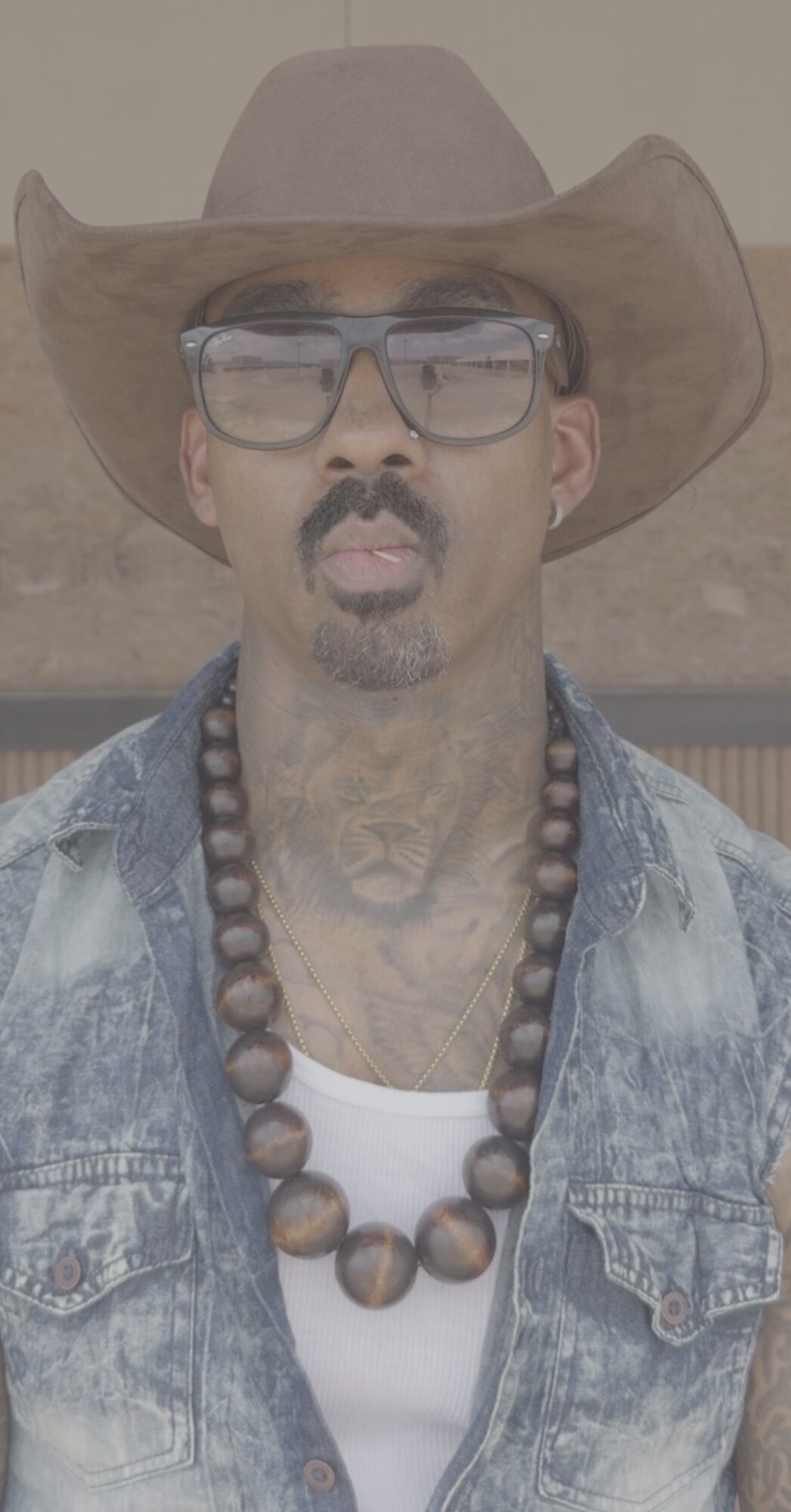 Man wearing a wide-brimmed hat, glasses, denim jacket, and beaded necklace, standing outdoors.